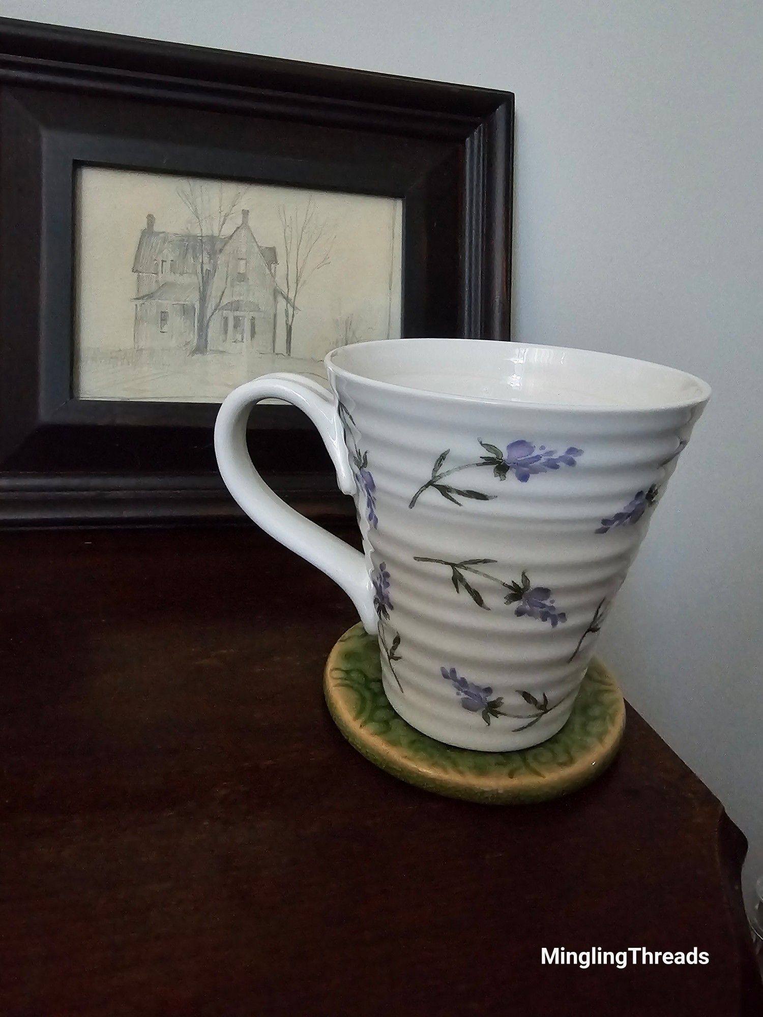 A dark wood side table. A cream coloured mug with a hand painted lavender sprig design. The mug us on a green pottery coaster. In the background is a dark wood frame is a pencil sketch of a lovely old house