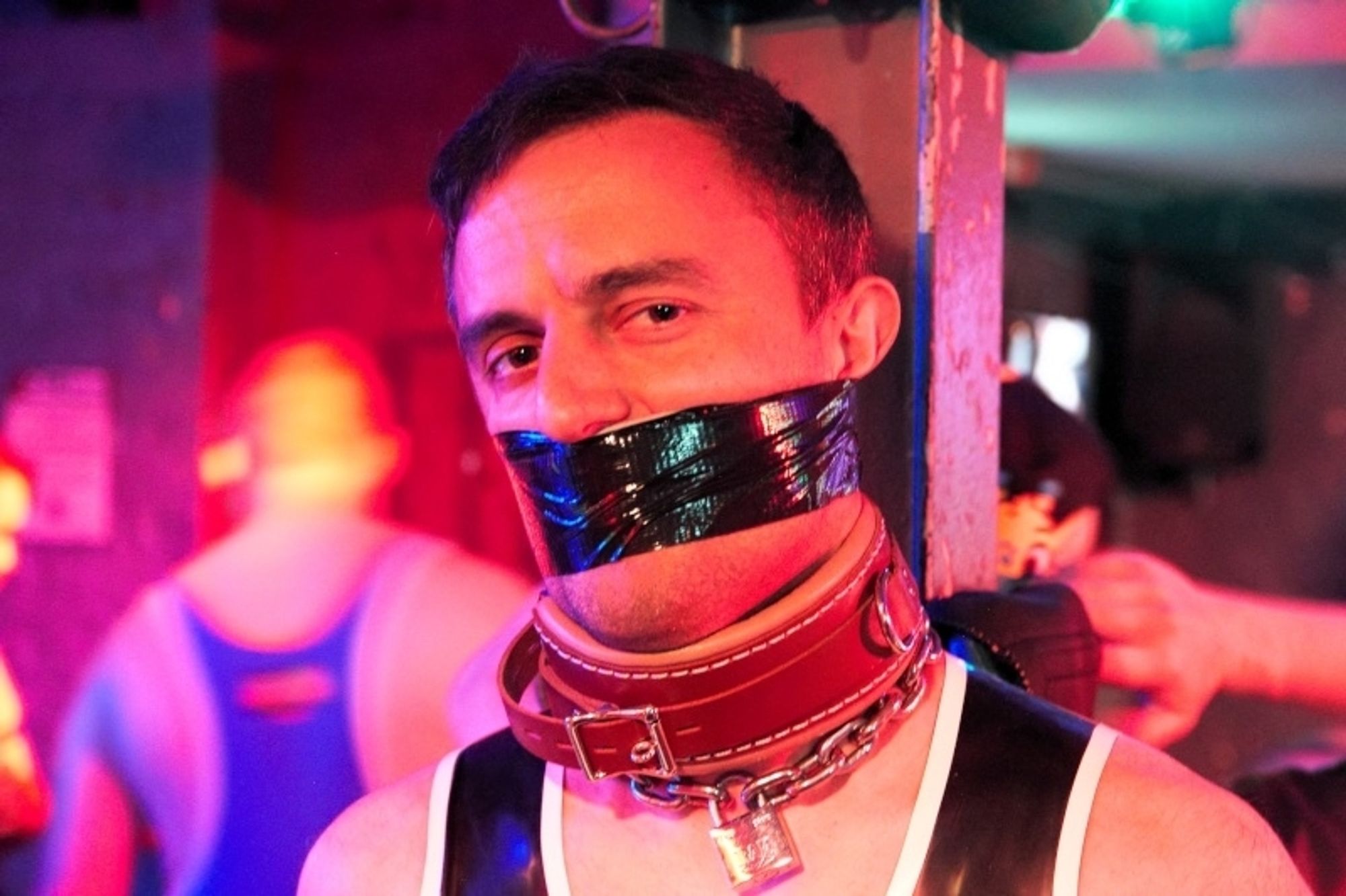 A guy tied to a column, with a tape gag and leather collar