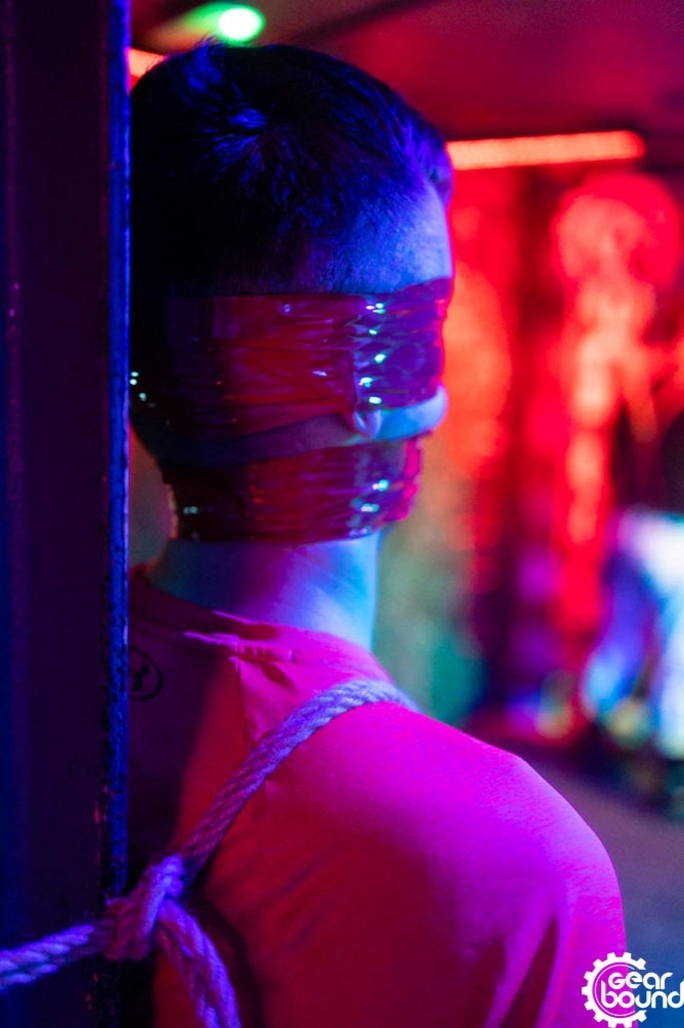 A rear view of a guy's head and shoulders. He is tied to a column with rope, and he has a red tape gag and blindfold.