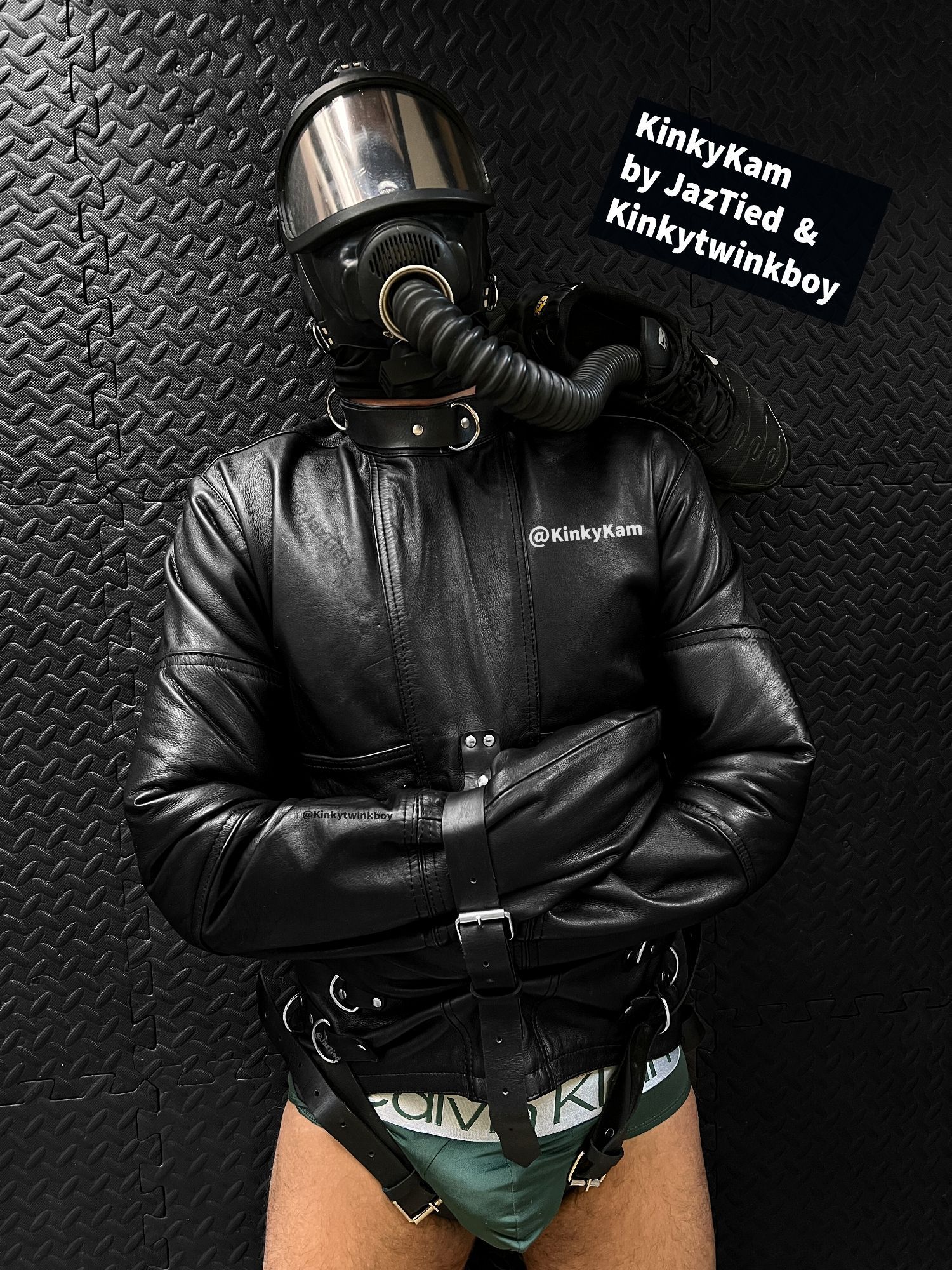 A guy wearing a pair of green briefs, restrained in a black leather straitjacket on the floor. He has a gas mask on, with the breathing tube connected inside a trainer.