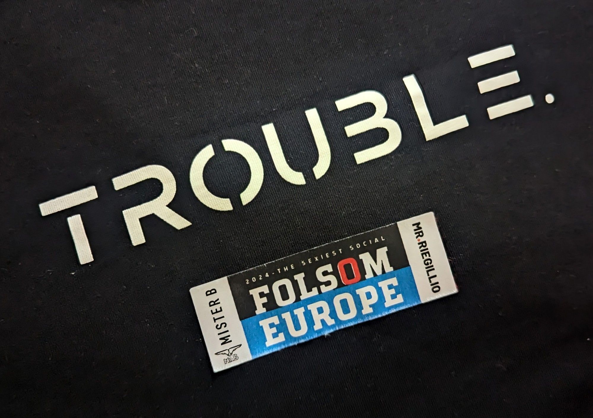 'Trouble' written in white text on a black t-shirt, with a Folsom sticker underneath