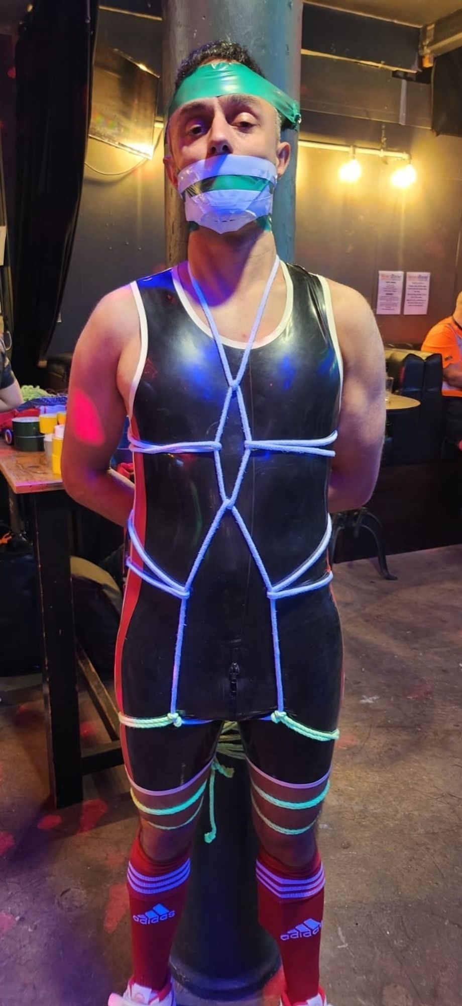 A guy tied to a column, wearing a black rubber singlet and red football socks. He is tied with a white rope body harness and his hands behind his back, and gagged with tape.