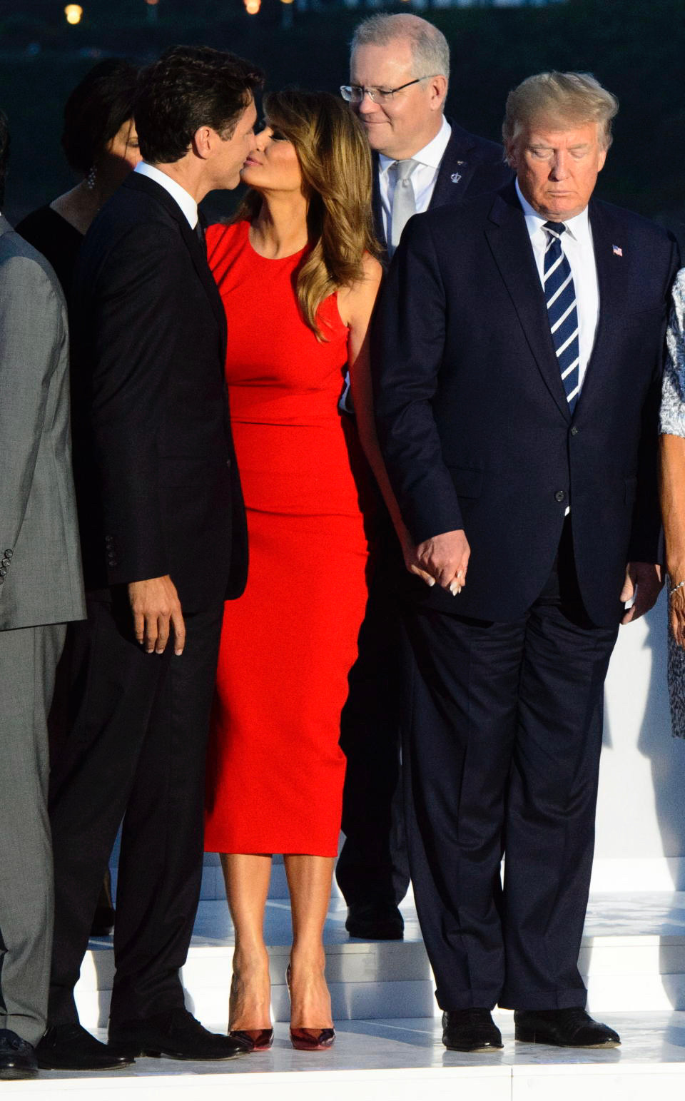 Press photo of Trudeau and Melania giving each other a cheek kiss.  She is smiling yet holding Trumps hand as he scowls and looks down.  The men are wearing dark suits. She is wearing a bright red body con sleeveless dress. They are standing on a stage