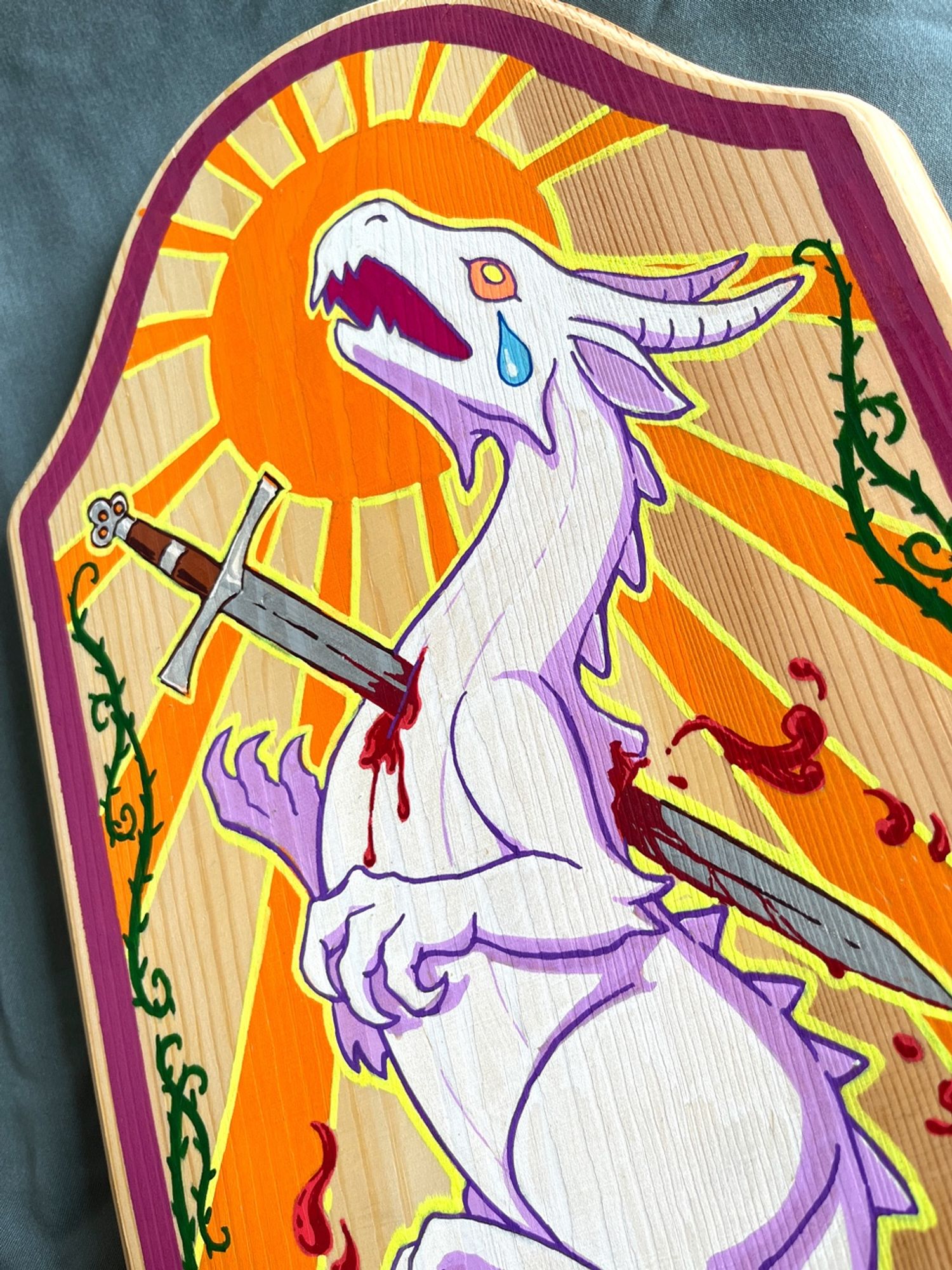 Close up photo of tall wooden plaque with painting of a heraldic-esque white dragon with a longsword plunging through its chest, bleeding, and with cut wings falling to the sides. The backdrop features an orange sun with long beams and ornate dark thorny vines twisting from the bottom up the sides. All within a raspberry colored frame.