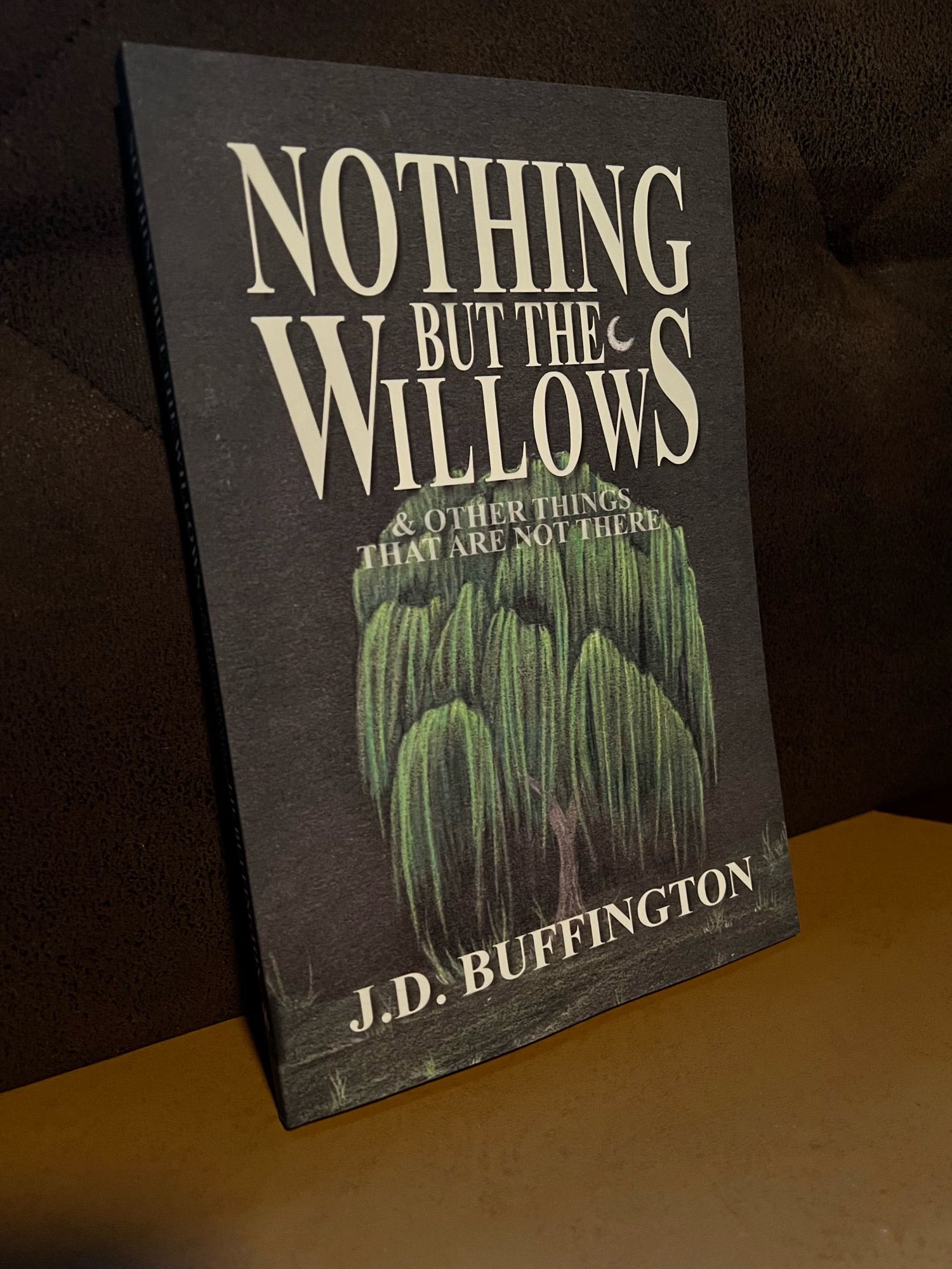 The paperback of Nothing But The Willows & Other Things That Are Not There by J.D. Buffington, featuring cover art by Stina Patton. The title is in large white font for the main title and a translucent grey for the subtitle. A mournful faced willow tree centers the cover in a nighttime scene with a crescent moon among the title letters.