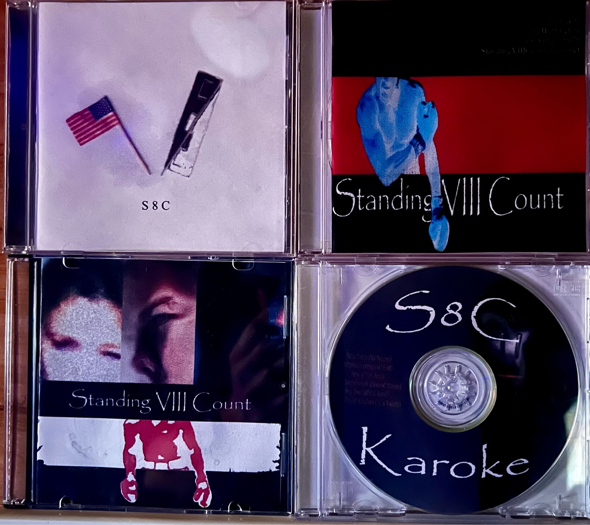 Album covers for the demo CDs JD Buffington made through the years for Standing VIII Count, the band. In the upper left is a mostly white cover with two small flags (an American flag toothpick, and an S8C banner made with staples and tape) and small “S8C” typed below. The upper right is striped top to bottom black, red, black with Muhammad Ali in negative color, appearing blue, over the red banner, “Standing VIII Count” in Papyrus is on the bottom black banner. The lower left is three obfuscated photos of members of the band, “Standing VIII Count” in Papyrus again, and another Ali silhouette in red over a white banner. The lower right is a black CD with “S8C” on top and “Karoke” on bottom in Papyrus.