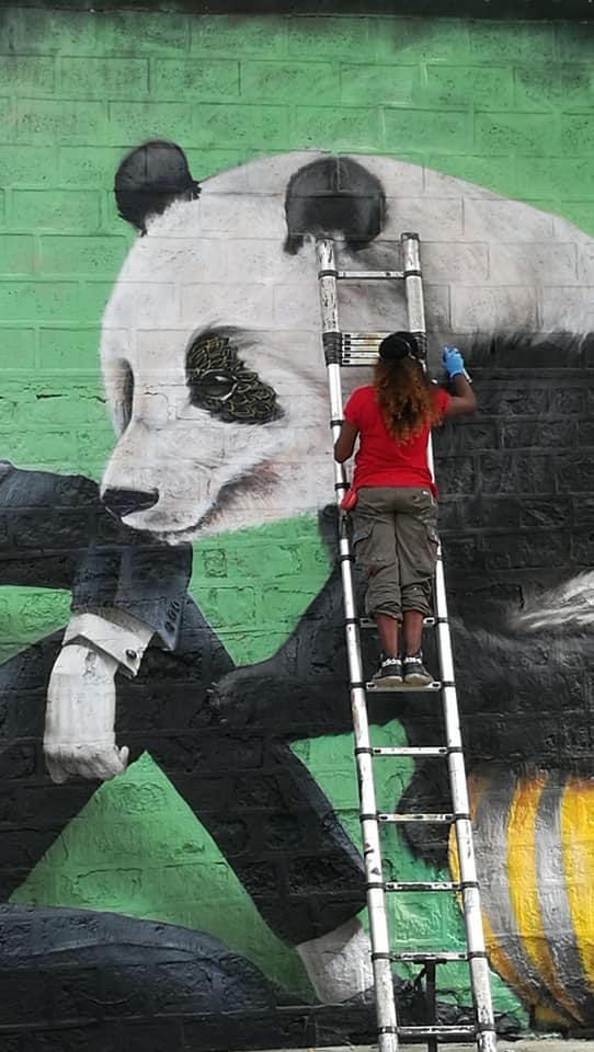 Street artiste perchée sur une échelle en train de mettre la touche finale à une grande fresque murale représentant un panda.