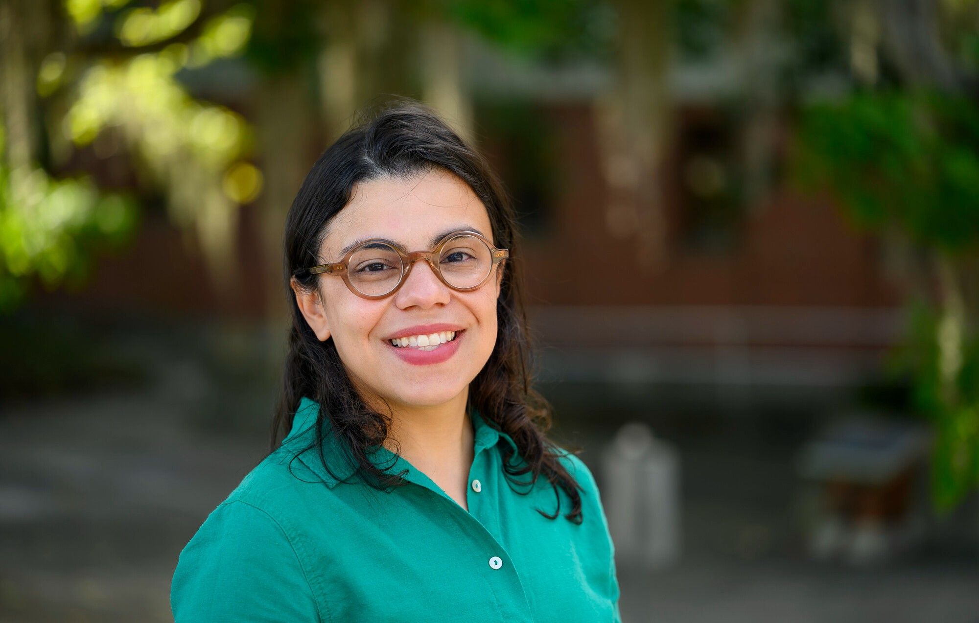 portrait of a person with glasses and dark hair smiling at the camera with a brick building and leaves out of focus in the background