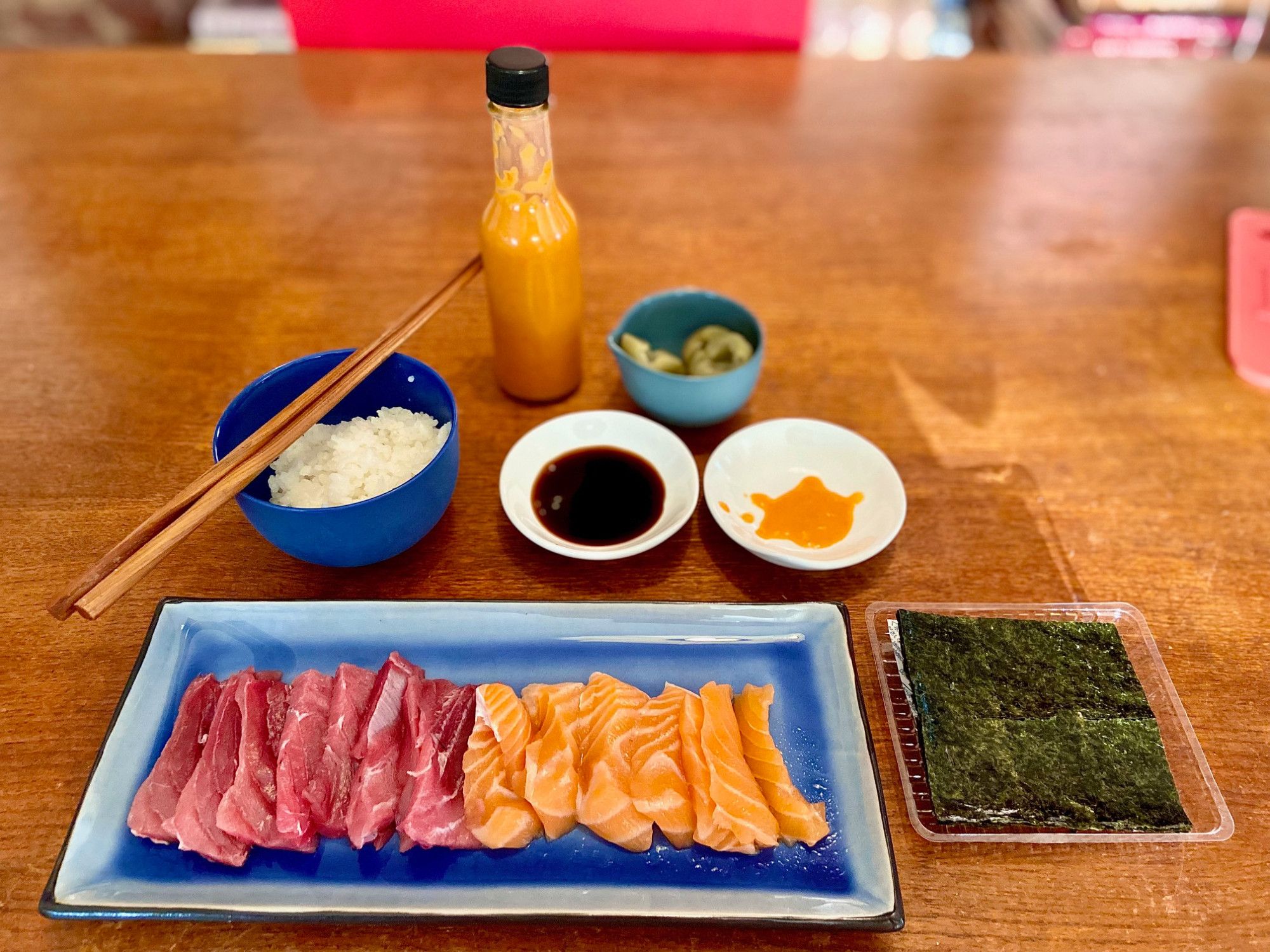 A blue rectangular plate with tuna and salmon slices of sashimi, a plastic tray of nori, a blue bowl of white rice and chopsticks, a small plate of soy sauce, small plate of hot sauce/bottle, and a blue bowl of Chinese pickles.