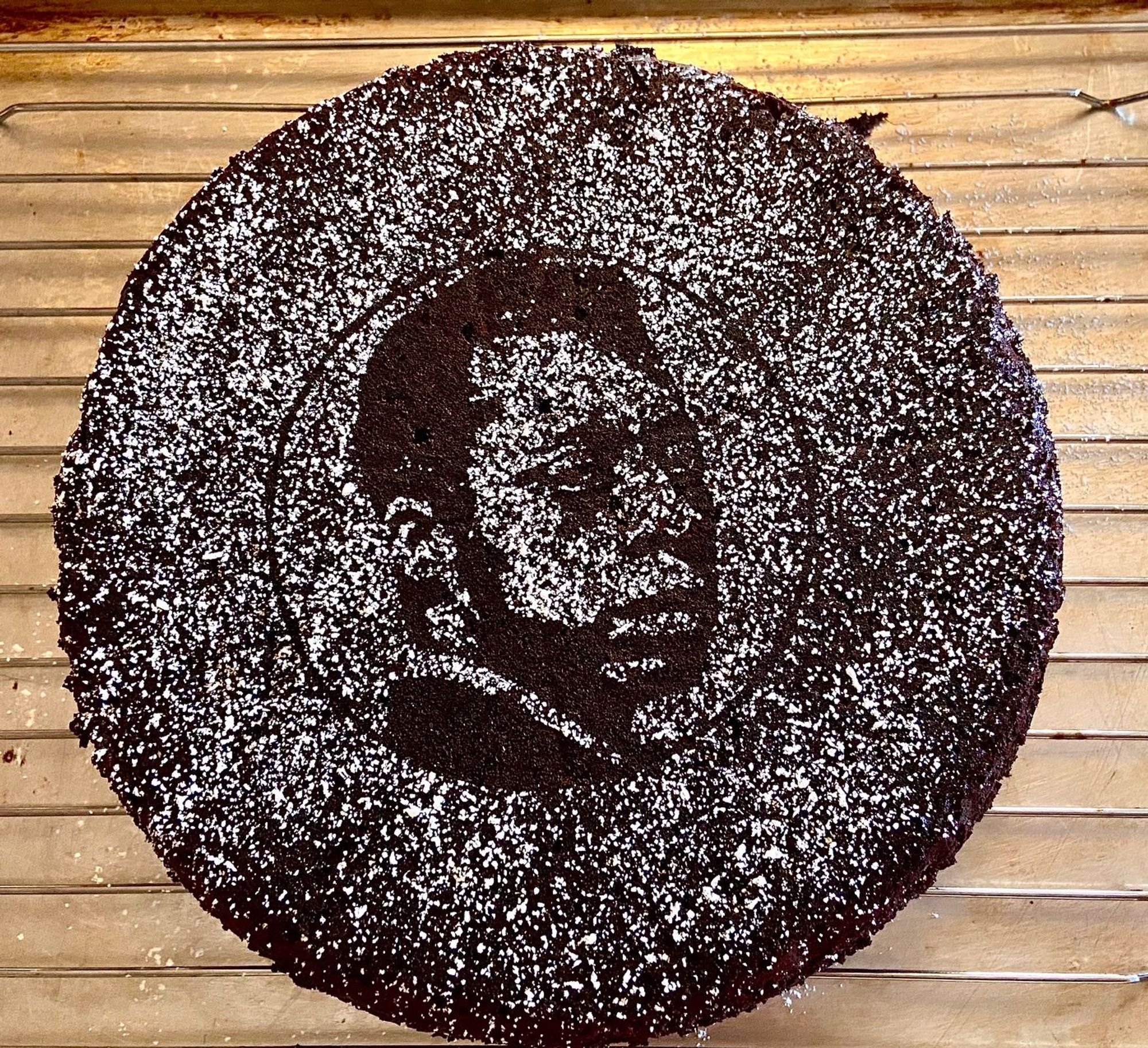 A round chocolate cake on a wire rack cooling. The cake's top is dusted with powdered sugar and has a portrait of James Baldwin created by a stencil.