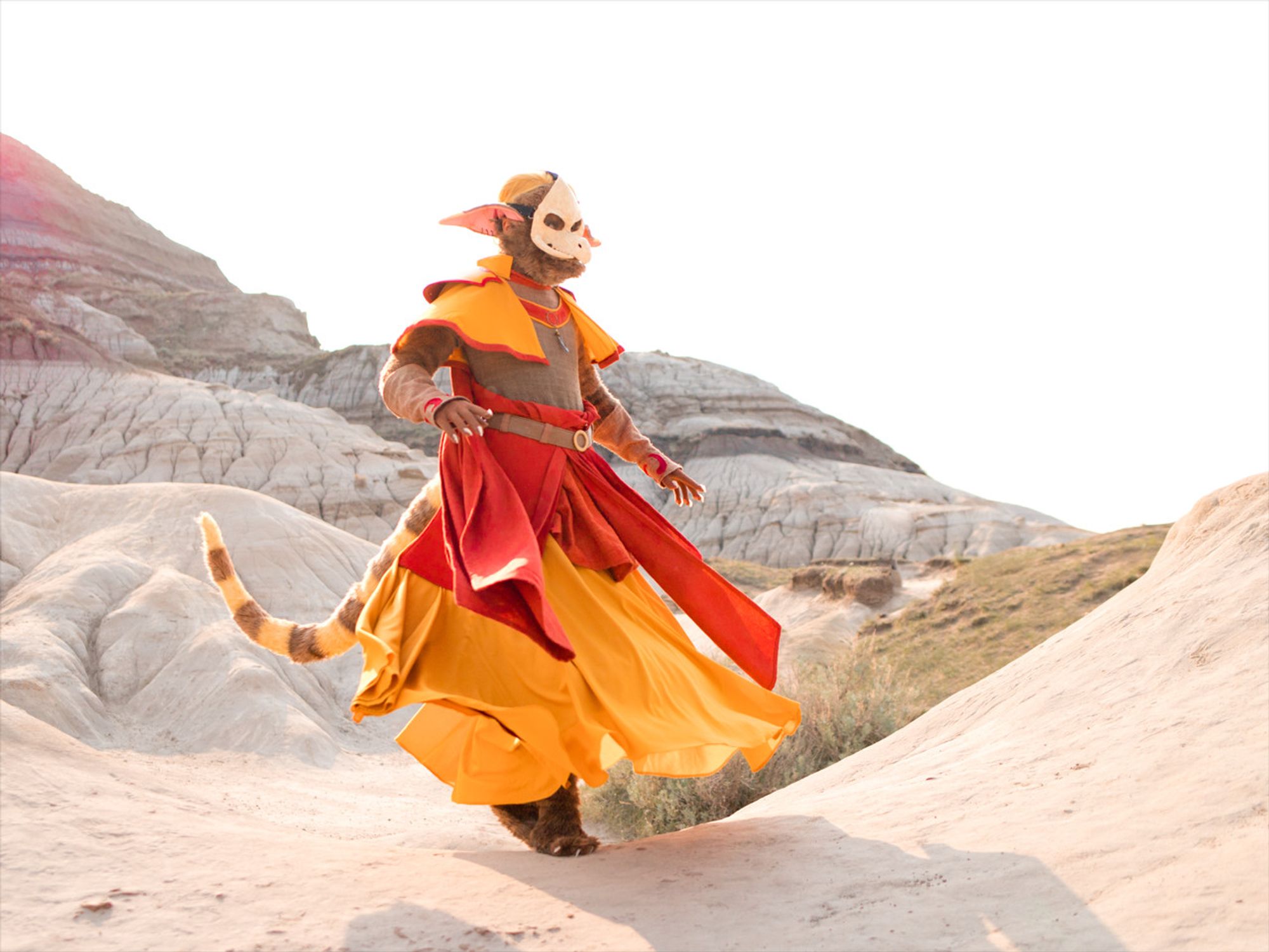 A photo of a costumer doing a spin, yellow and red fabric billowing out around him. He is dressed as an anthropomorphic character, with a long striped tail and ears. The setting is a mountainous desert region.