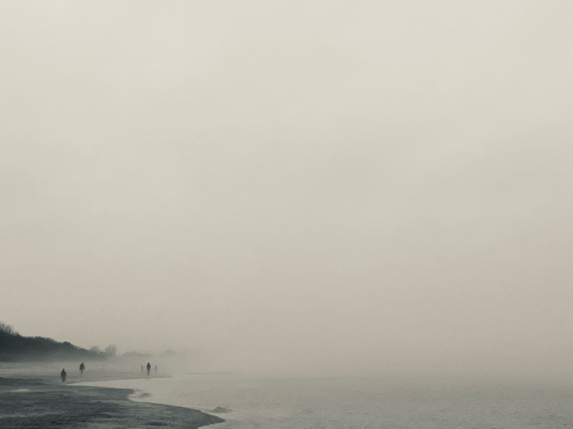 Nebel zieht über die Ostsee. Links im Bild sind etwas weiter entfernt am Strand schemenhaft 3 Personen zu erkennen, die in den Nebel laufen, der bereits 2 noch weiter entfernte Personen fast verschluckt hat. Im Bildhintergrund links ist von Nebel bedeckter Wald zu sehen, der Blick in die Ferne aufs Wasser wird in der Aufnahme in Grautönen vom Nebel verdeckt.