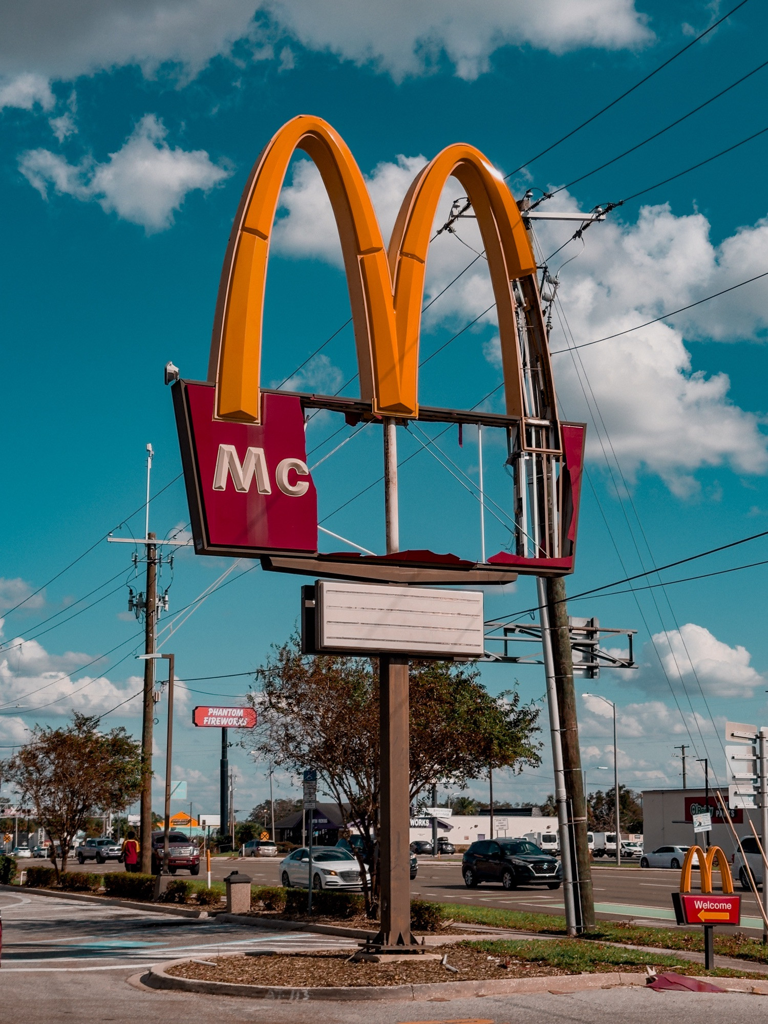 Damaged McDonald’s sign from the aftermath of Hurricane Milton. The iconic Golden Arches appear tattered, with parts missing, particularly on the maroon panel beneath the arches, which only displays “Mc,” evoking a sense of irony as if rebranded to “McMilton’s.”