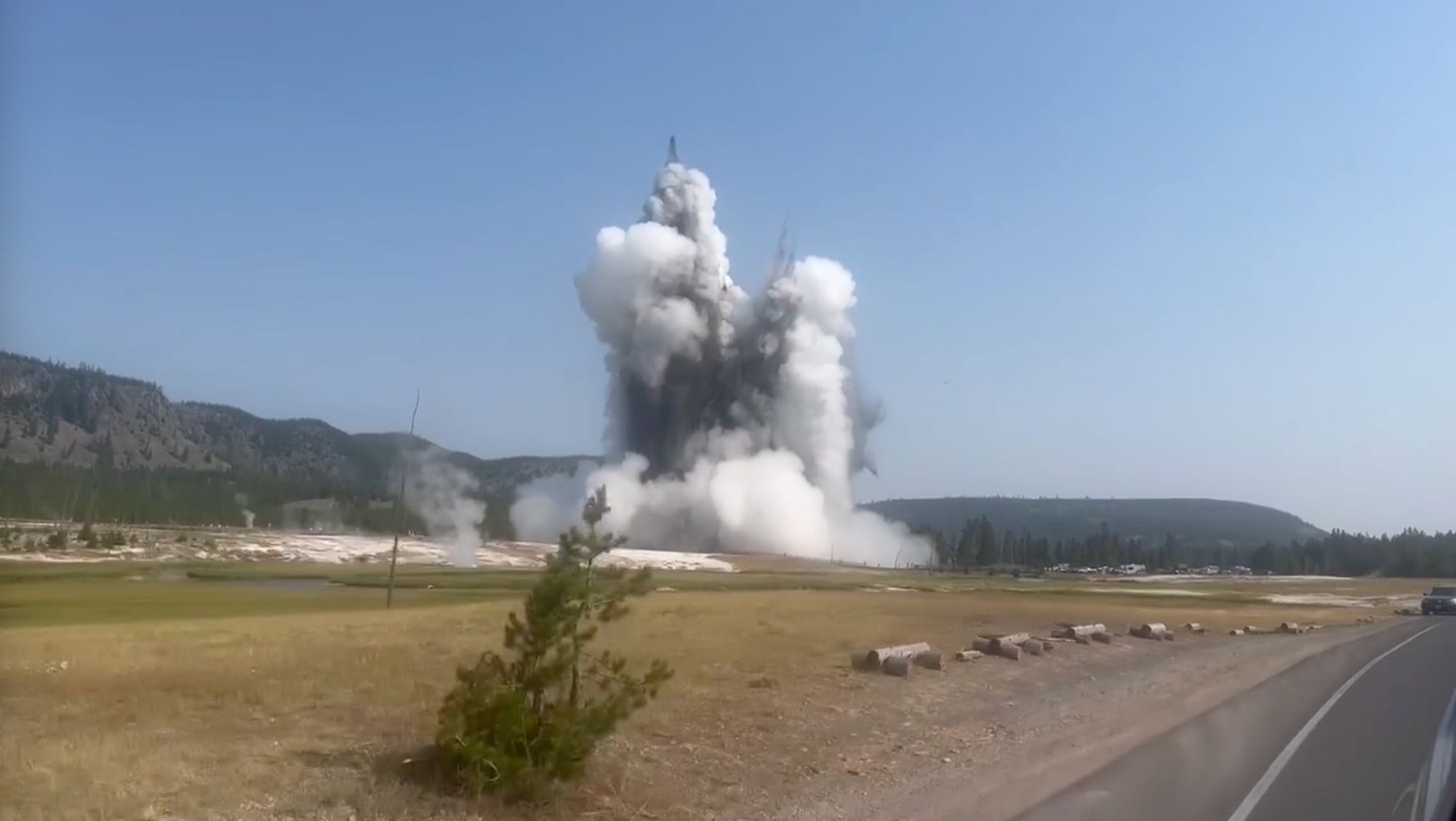 Large jets of black water contrasted against clouds of white steam erupt in the distance in this photo shot from the passenger side of a vehicle on a road.