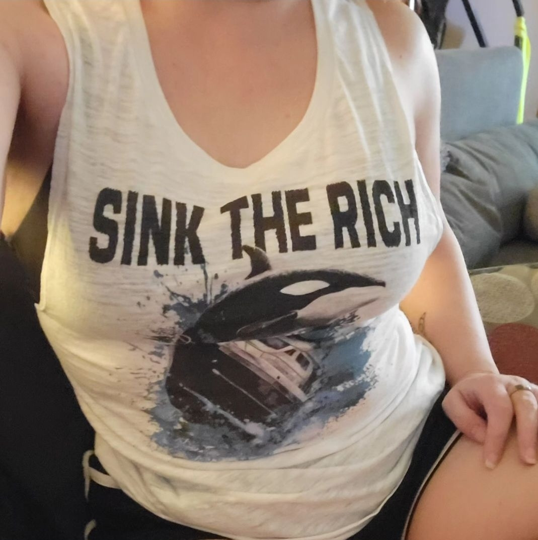 A torso shot of a woman wearing a tank top shirt. It's white and it has a blue and black and white image on the front of an orca tipping over a yacht. Above it reads "SINK THE RICH" in black lettering.