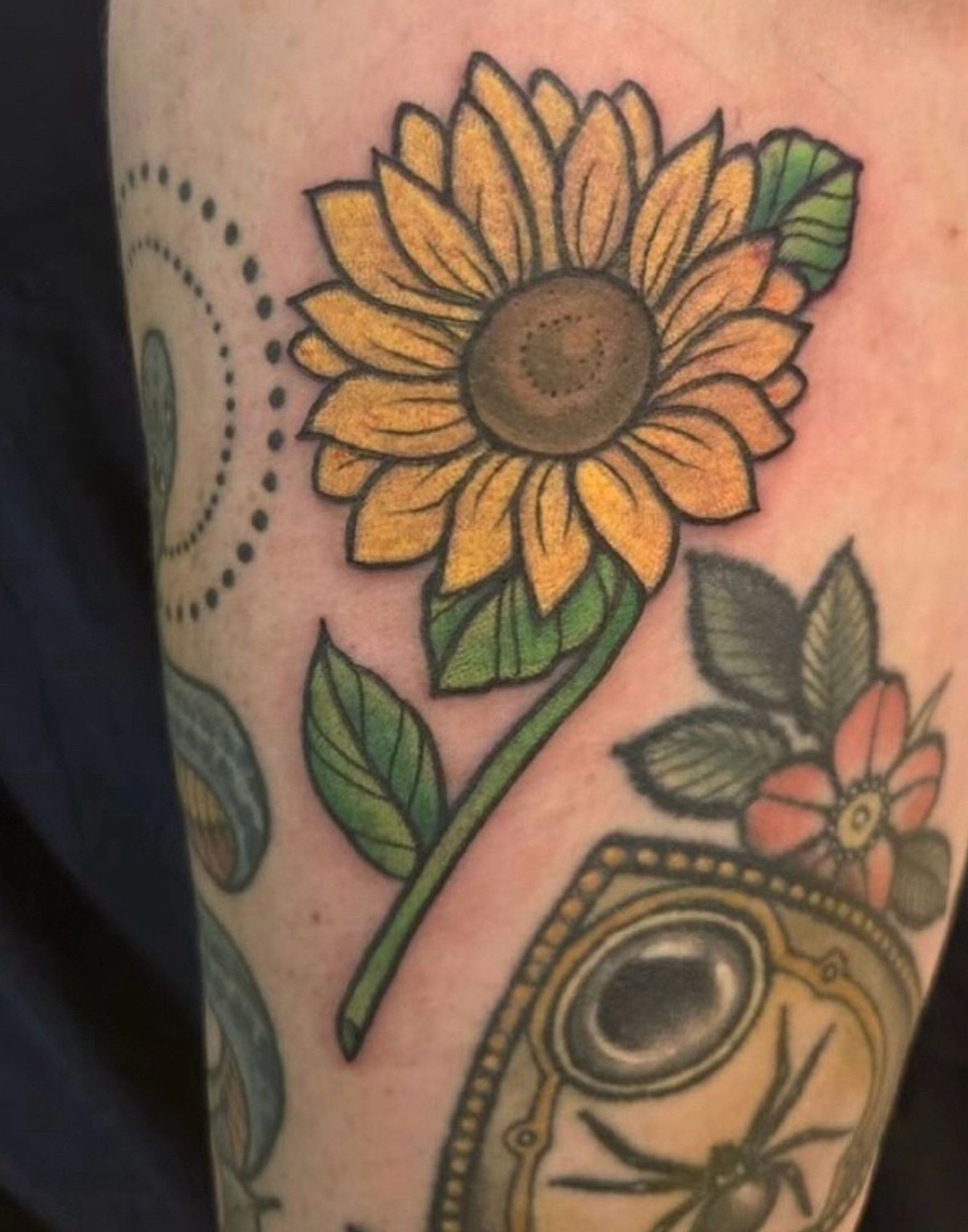 Closeup tattoo of a forearm with a sunflower with partial view of a snake tattoo and a ouija planchette tattoo with flowers and a spider