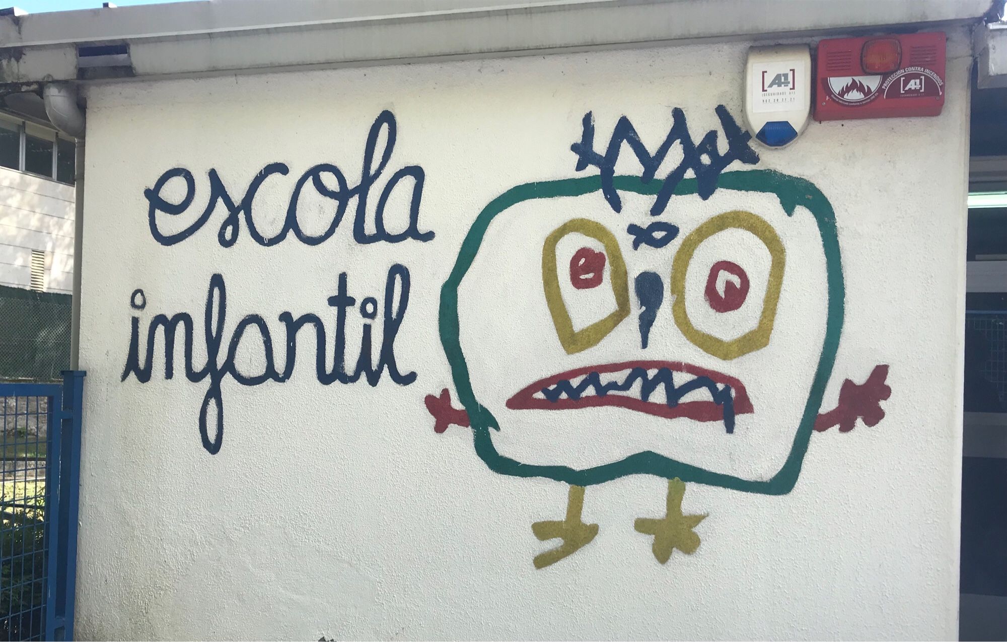 Muro con la inscripción “escola infantil”. Un horripilante y grotesco ser está dibujado al lado con trazos de colores.