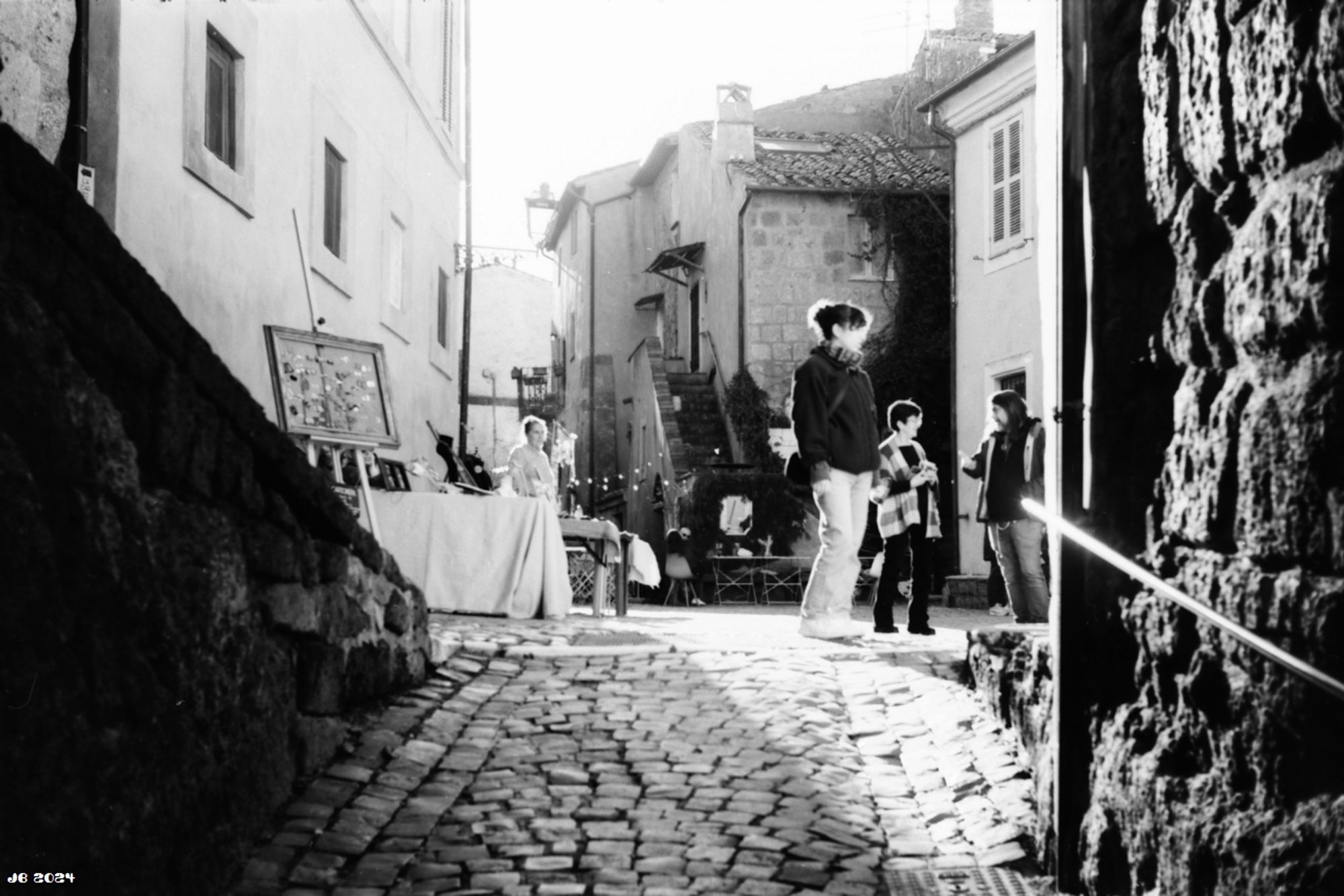 Schwarzweißfoto - Blick durch das Eingangstor in das Zentrum der kleinen historischen Stadt Calcata über Pflastersteine. Links und rechts Mauern und Häuser, in der Bildmitte ein kleiner Markt mit Kunstprodukten und verschiedene Leute - Aussteller und Besucher.