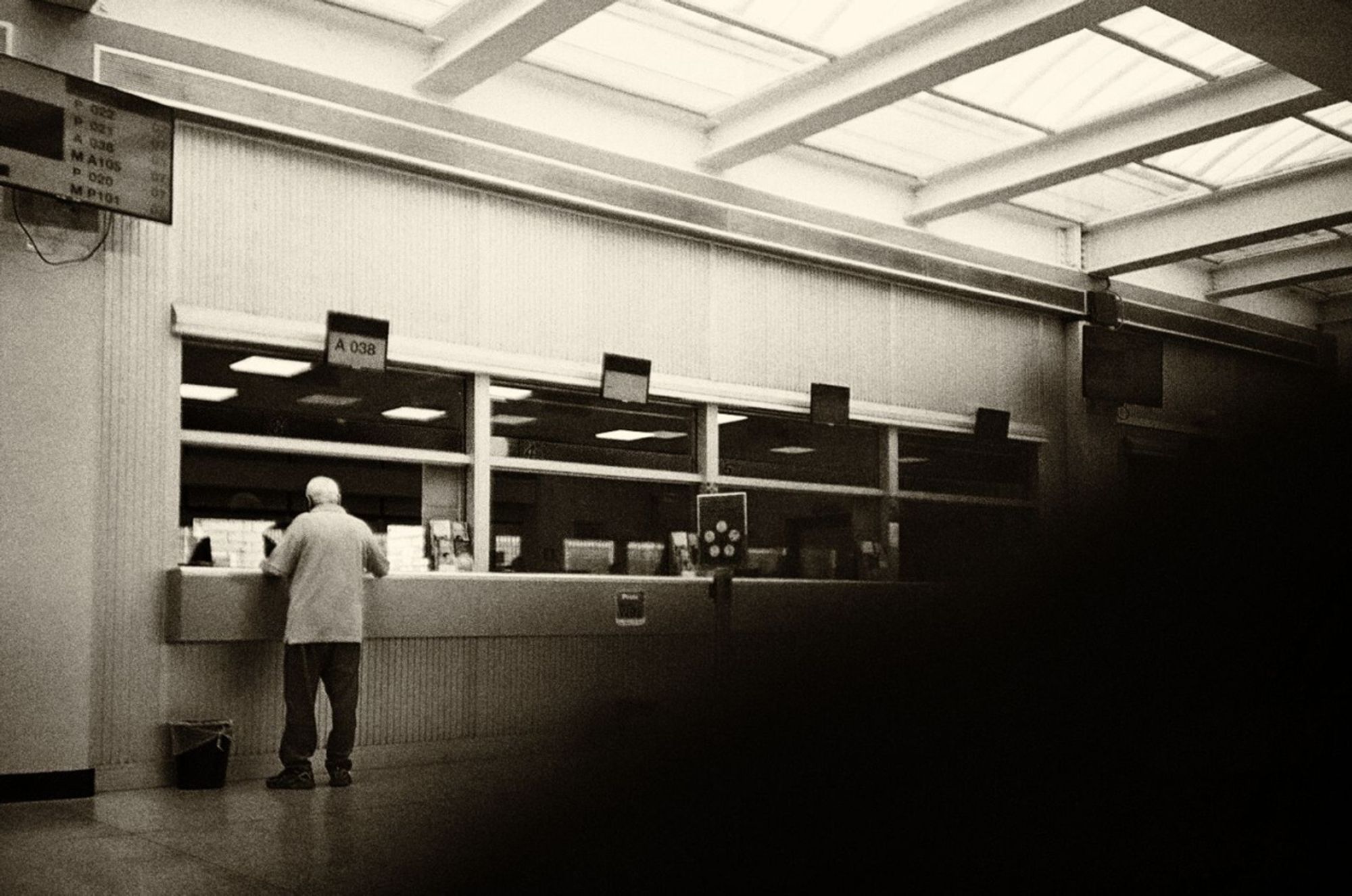 Monochrome photo taken inside a post office in Italy while waiting. There are four counters but there is only one person being served on the left. The bottom right of the photo is obscured by a finger in front of the lense. Lomo LC-A on Kodak Gold film, desaturated and edited.