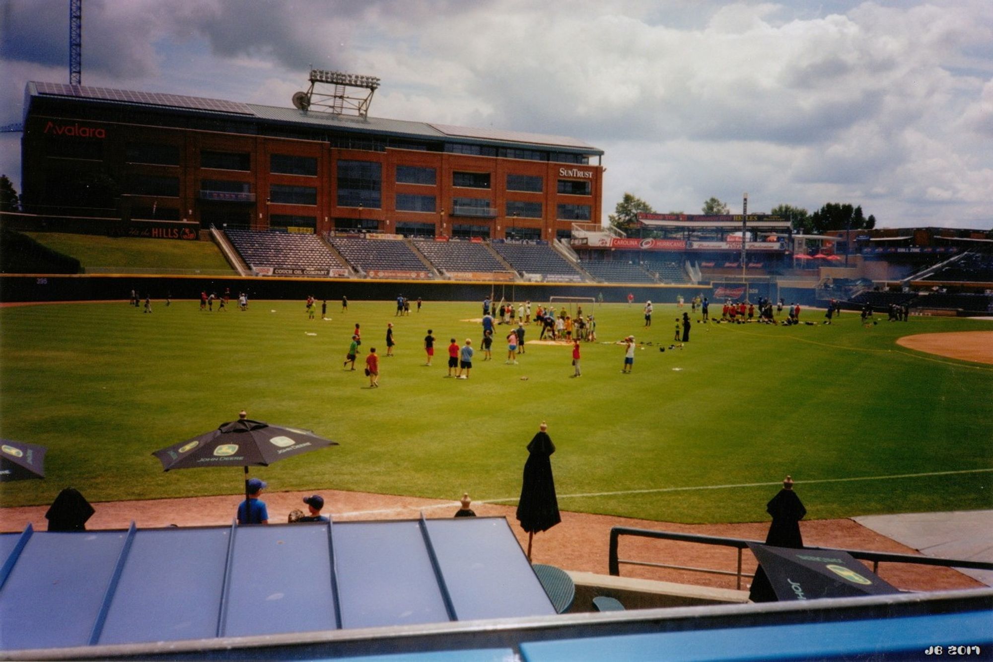 Farbfoto - ein Teil des Baseball-Stadions der Durham Bulls in Durham, North Carolina, USA. In der Bildmitte grüner Rasen und Leute, im Hintergrund Teile der Zuschauertribüne, dahinter ein großes braunes Backsteingebäude. Bewölkter Himmel.