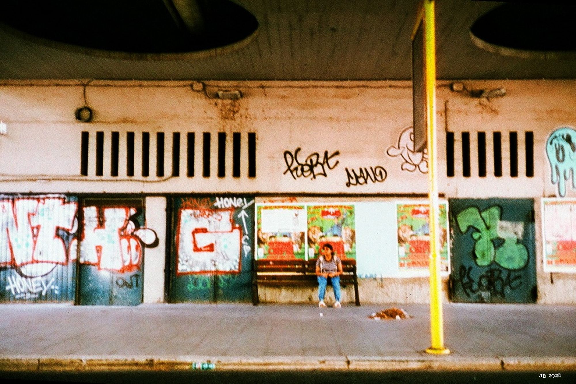 A young woman waiting for the bus on a bench at a bus stop in Rome. The wall of the building behind her is full of big graffiti in all colors.