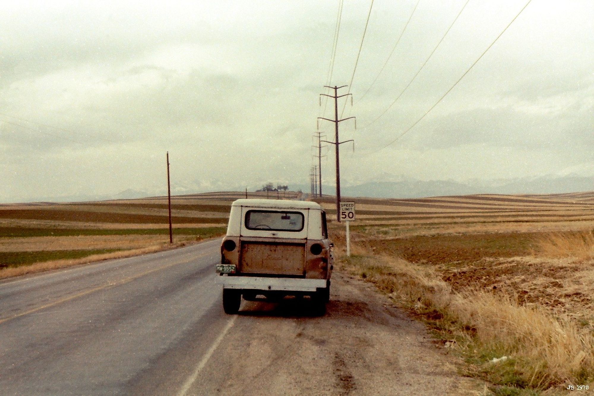 Altes Farbfoto von 1978 - mein 1966er International Scout hatte unterwegs in Colorado den Geist aufgegeben, so dass er am Straßenrand in der kargen Landschaft steht. Rechts und links Äcker und Stoppelfelder. An der Straße entlang Strommasten und Leitungen. Bedeckter grauer Himmel.