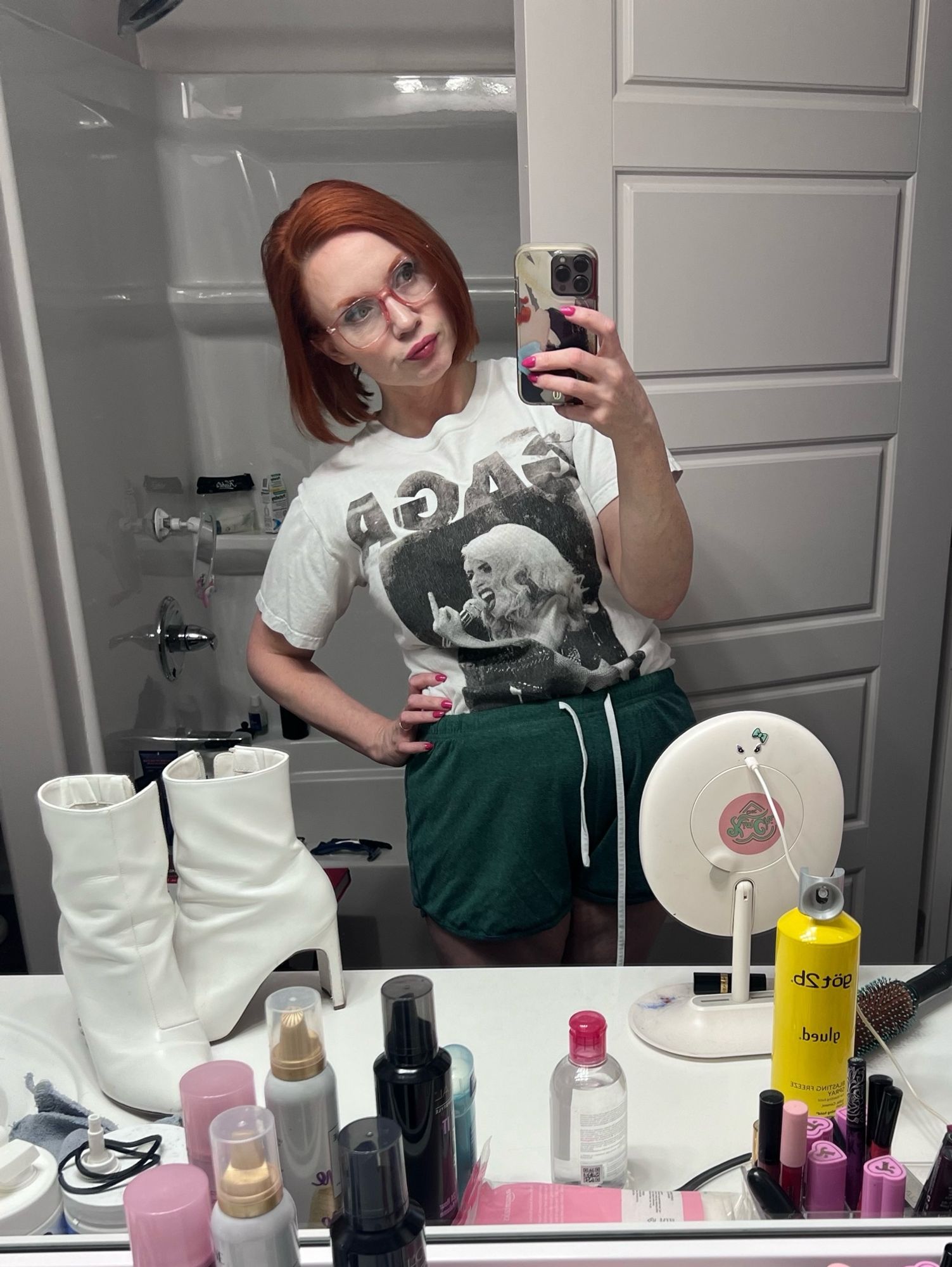 Mirror selfie of a cluttered bathroom counter and white woman with red hair wearing a white Lady Gaga tshirt and green shorts.