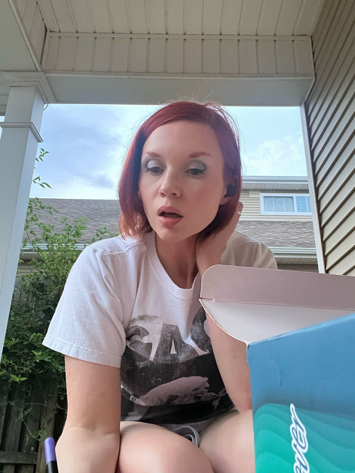 White woman with red hair on a patio in Lady Gaga tshirt and shorts with a box of markers in the foreground.