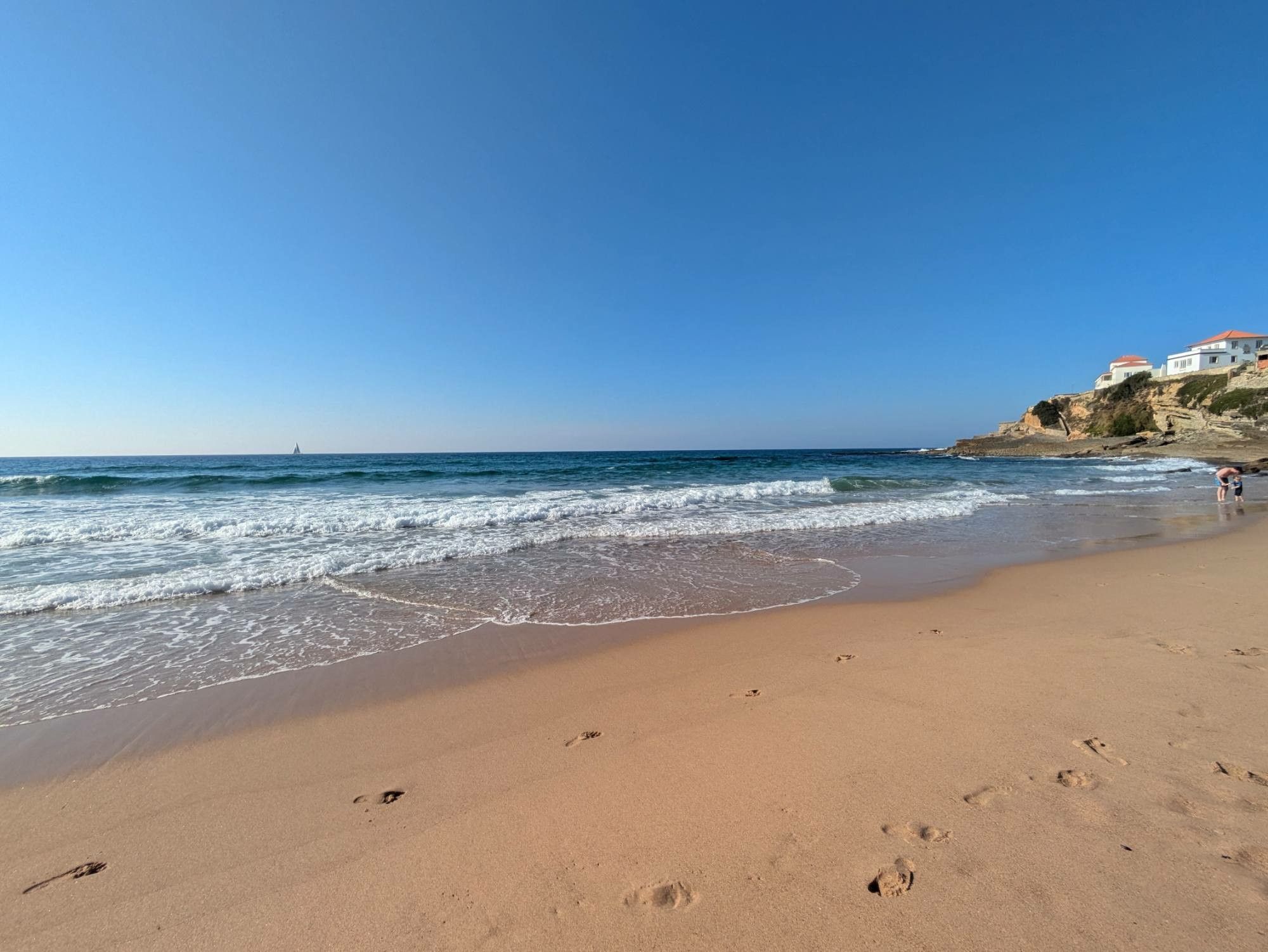 im Vordergrund Sandstrand, dann weiße Wellenkämme und blaugrünes Meer dahinter. Ein paar Häuser auf Felsen am rechten Rand. Blauer wolkenloser Himmel 