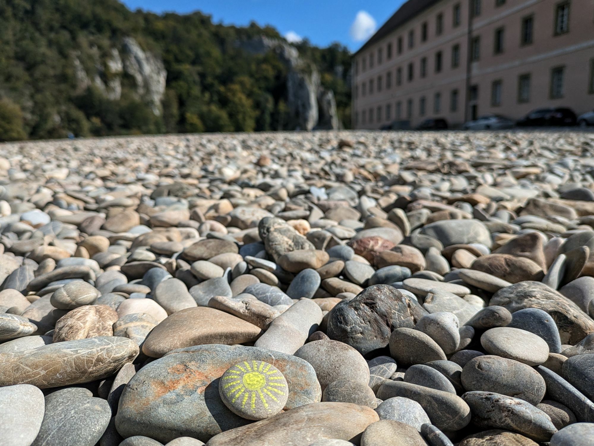 ein mit einer Sonne bemalter Stein liegt zwischen vielen Kieseln.
Rechts ist ein Flügel vom Kloster und links steiler Fels