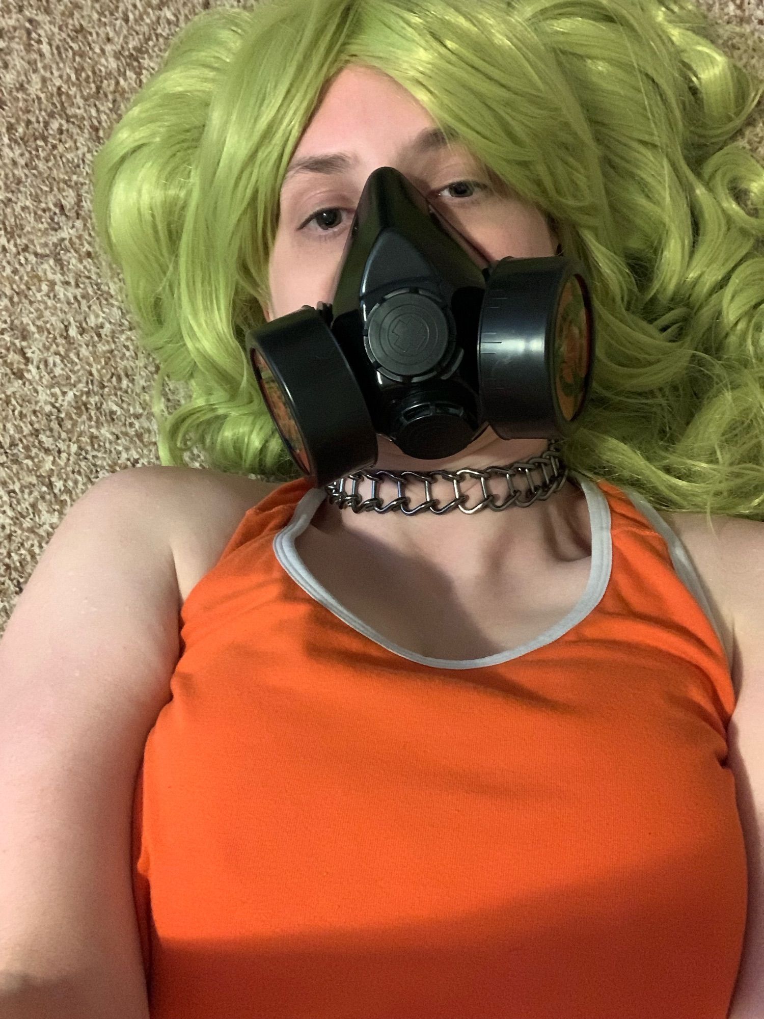 Female bodied individual lying down on her back in a bright green wig wearing a face mask, choker necklace and bright orange sports bra.