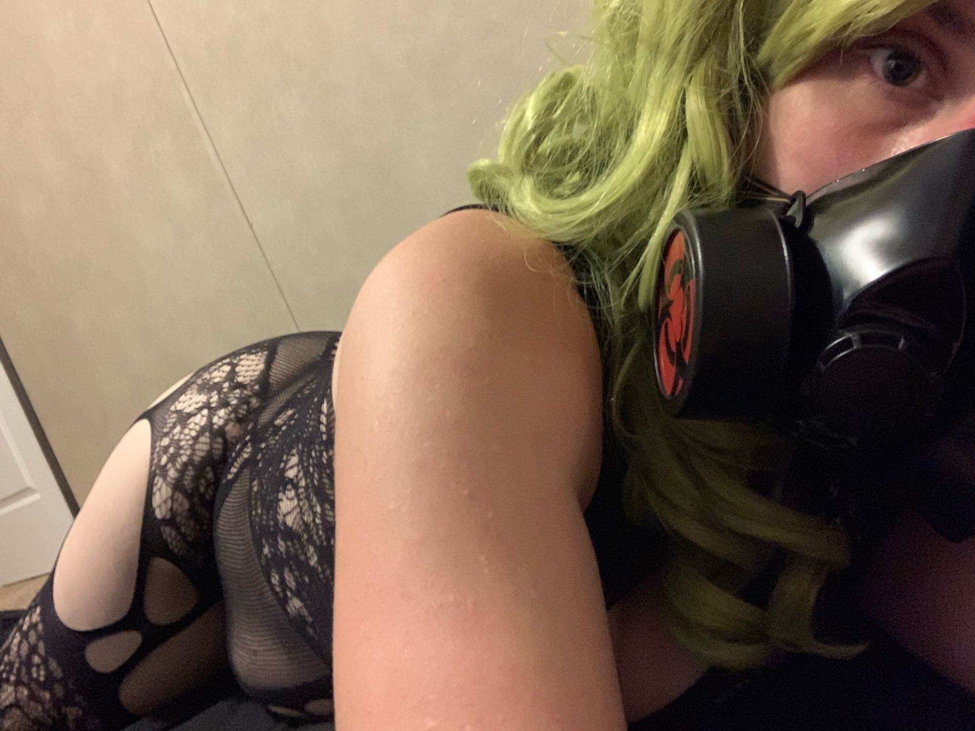 Female bodied person with bright green hair lying on her side, looking at the camera while wearing a mask and revealing lingerie.