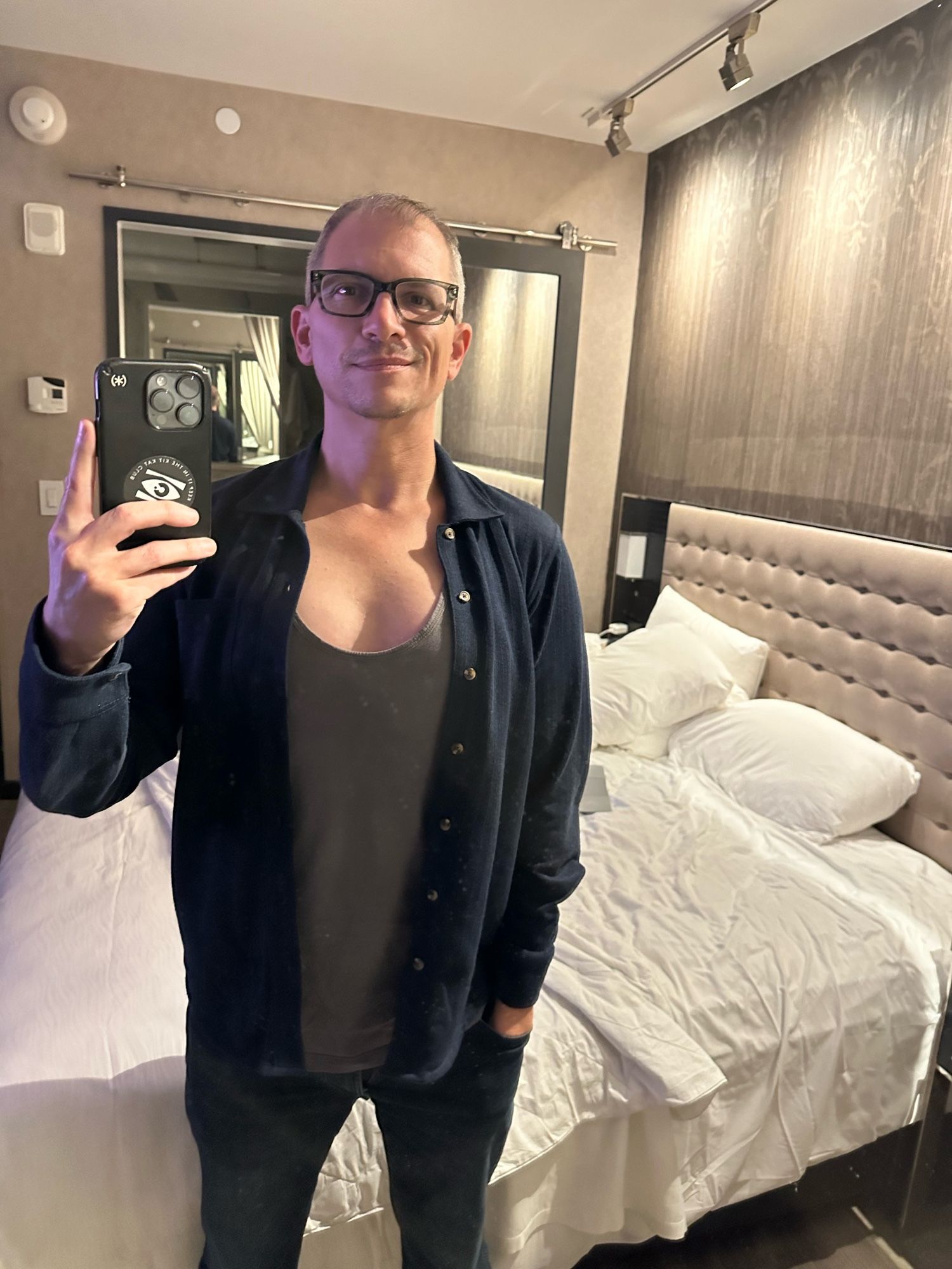 Chris in a grey tank and unbuttoned dark blue long-sleeved shirt and jeans in a hotel room.