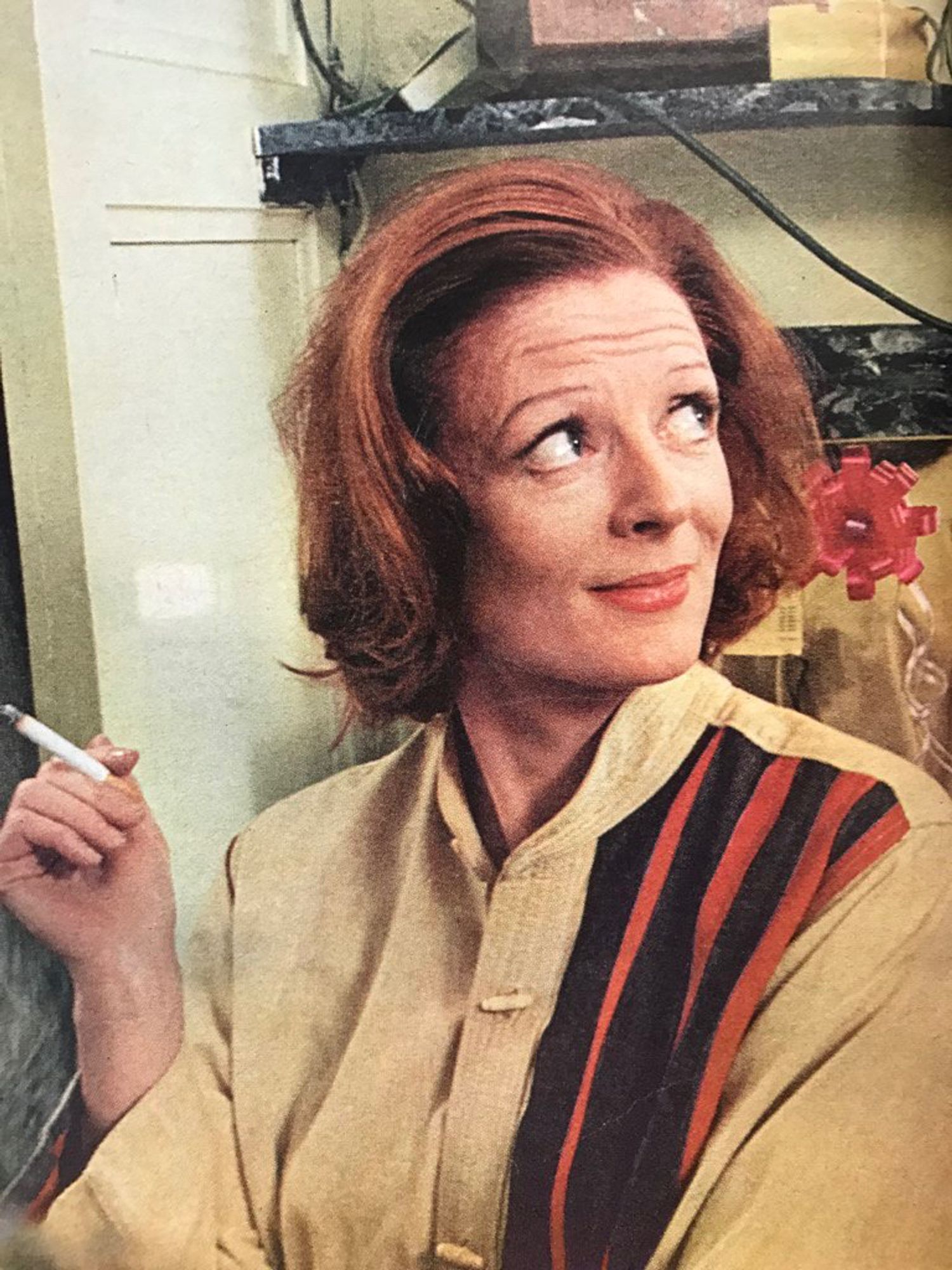 Maggie Smith with chin-length red hair, wearing a khaki jacket with orange and black stripes on the left half o the front of it. She's looking wryly up and back over her left shoulder at something or someone out of frame while she holds a cigarette in her right hand