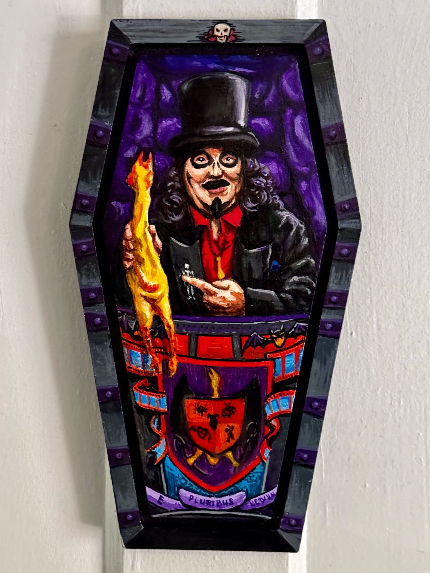 A coffin shaped wooden panel painted to look like Chicago-area TV horror host Svengoolie inside his signature coffin. He’s a chubby white guy with a curly black wig and a painted-on mustache and goatee as well as black rings around his eyes. He’s wearing a top hat and a black jacket over a red button up shirt. He pointing at the rubber chicken he’s holding in his opposite hand. The front of the lower half of the coffin is decorated with a rubber chicken coat of arms motif.