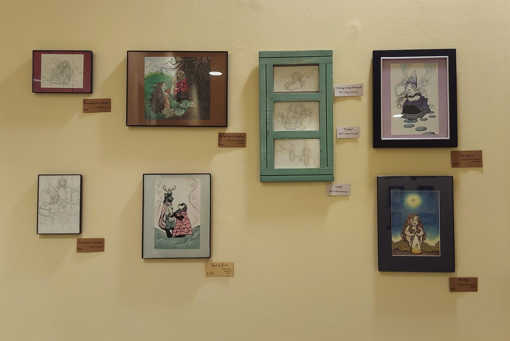 A photograph of a gallery wall, with a grid of 7 frames (one frame containing 3 pieces together), all featuring artwork of various faerie and fantasy characters.