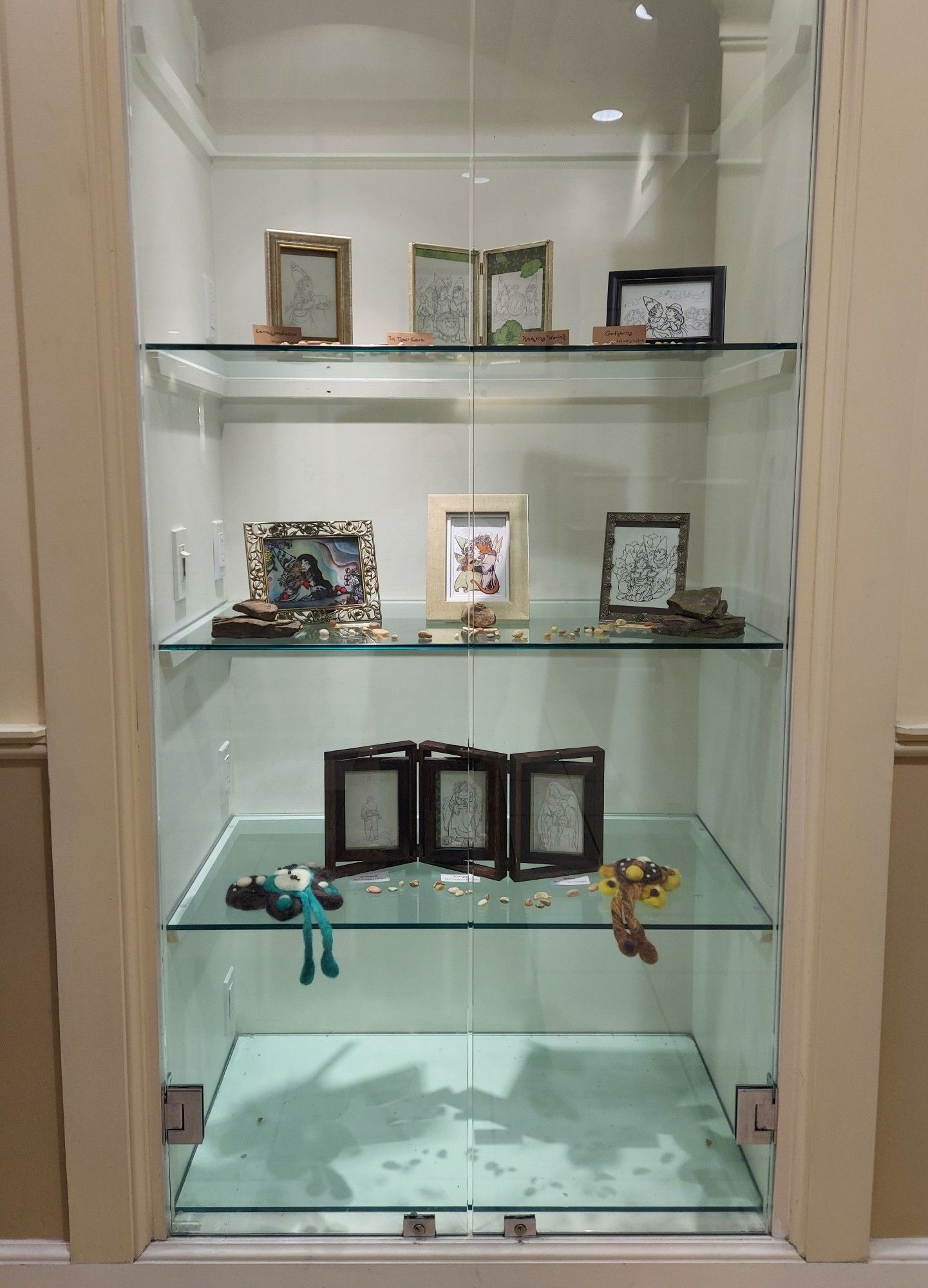 A photograph of a glass case with three shelves, displaying several framed pieces of art and two needlefelted pieces, all artwork featuring various faerie and fantasy characters.