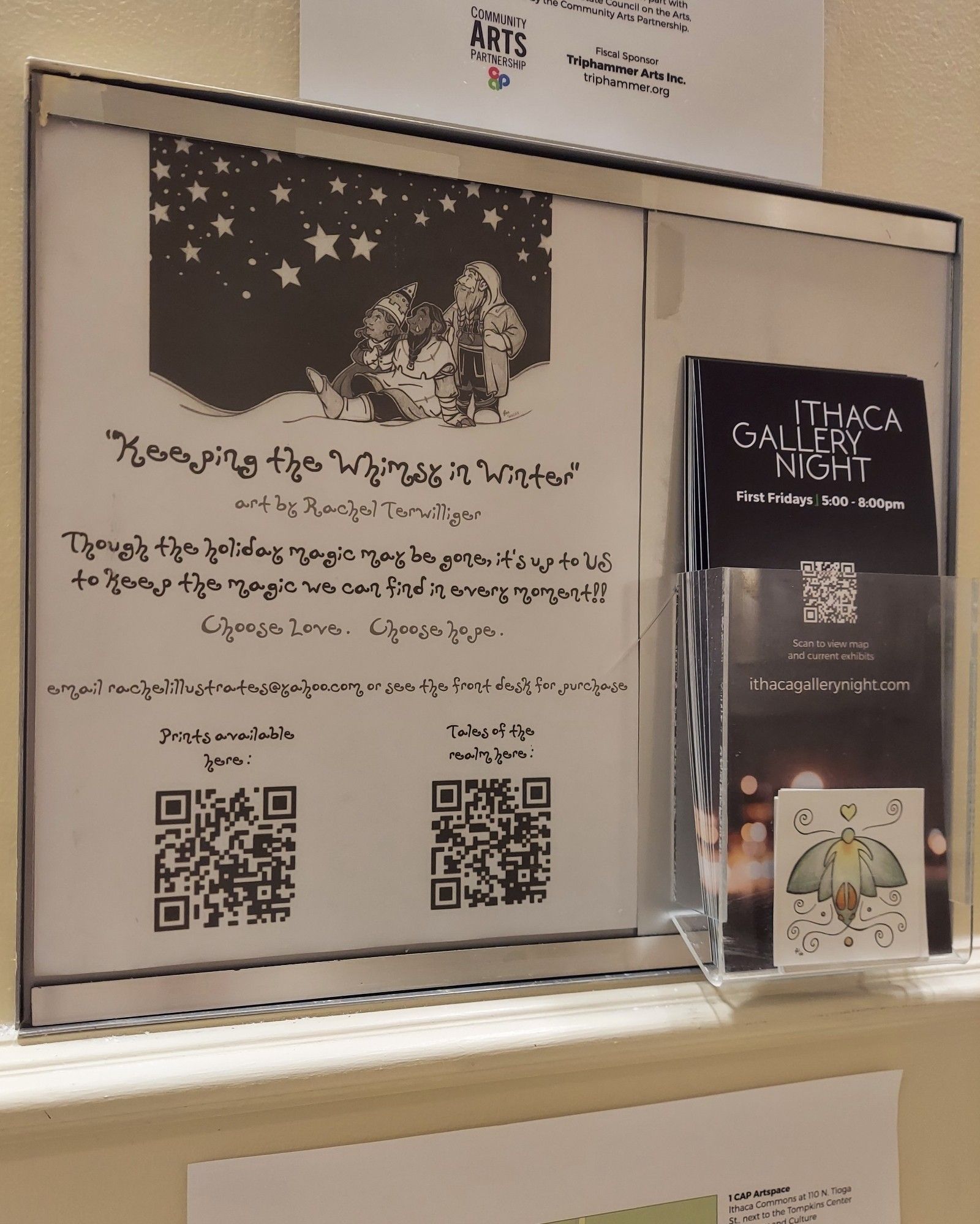 A photograph of the sign advertising and describing an art show. At the top of the page there is a drawing of three faerie beings looking up at stars in a winter landscape, with the text below: ” “Keeping the Whimsy in Winter” – art by Rachel Terwilliger – Though the holiday magic may be gone, it’s up to US to keep the magic we can find in every moment!! – email rachelillustrates@yahoo.com or see the front desk for purchase – Prints available here: (with QR code below) – Tales of the realm here: (with QR code below).”