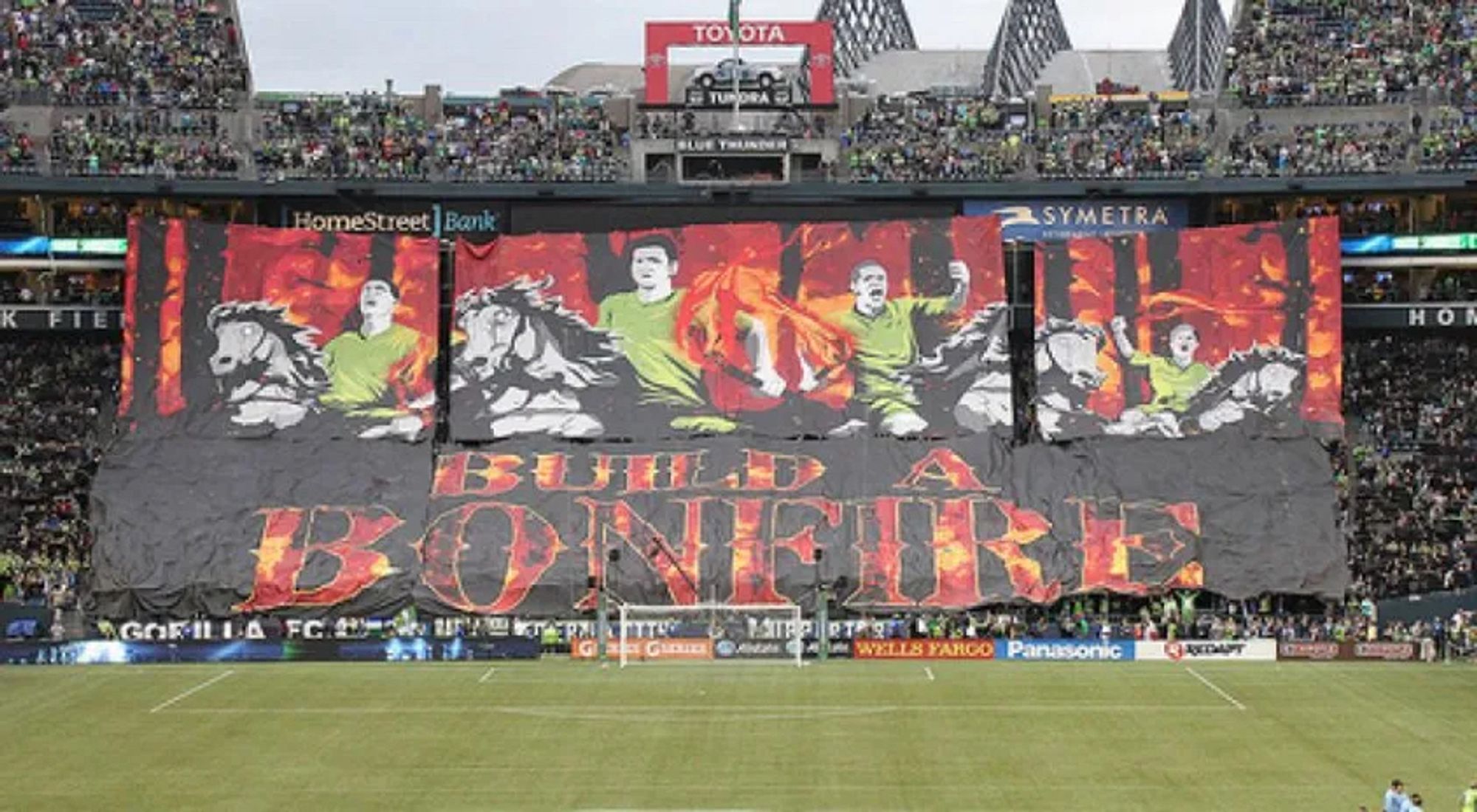 Emerald City Supporters 2013 tifo, a 40,000 sq ft masterpiece in depicting players in green riding black and white horses out of a forest consumed in red and orange flame, in multiple panels hanging down from the 3rd level of a stadium. Below a large banner reads Build a Bonfire in flaming letters on a black background.