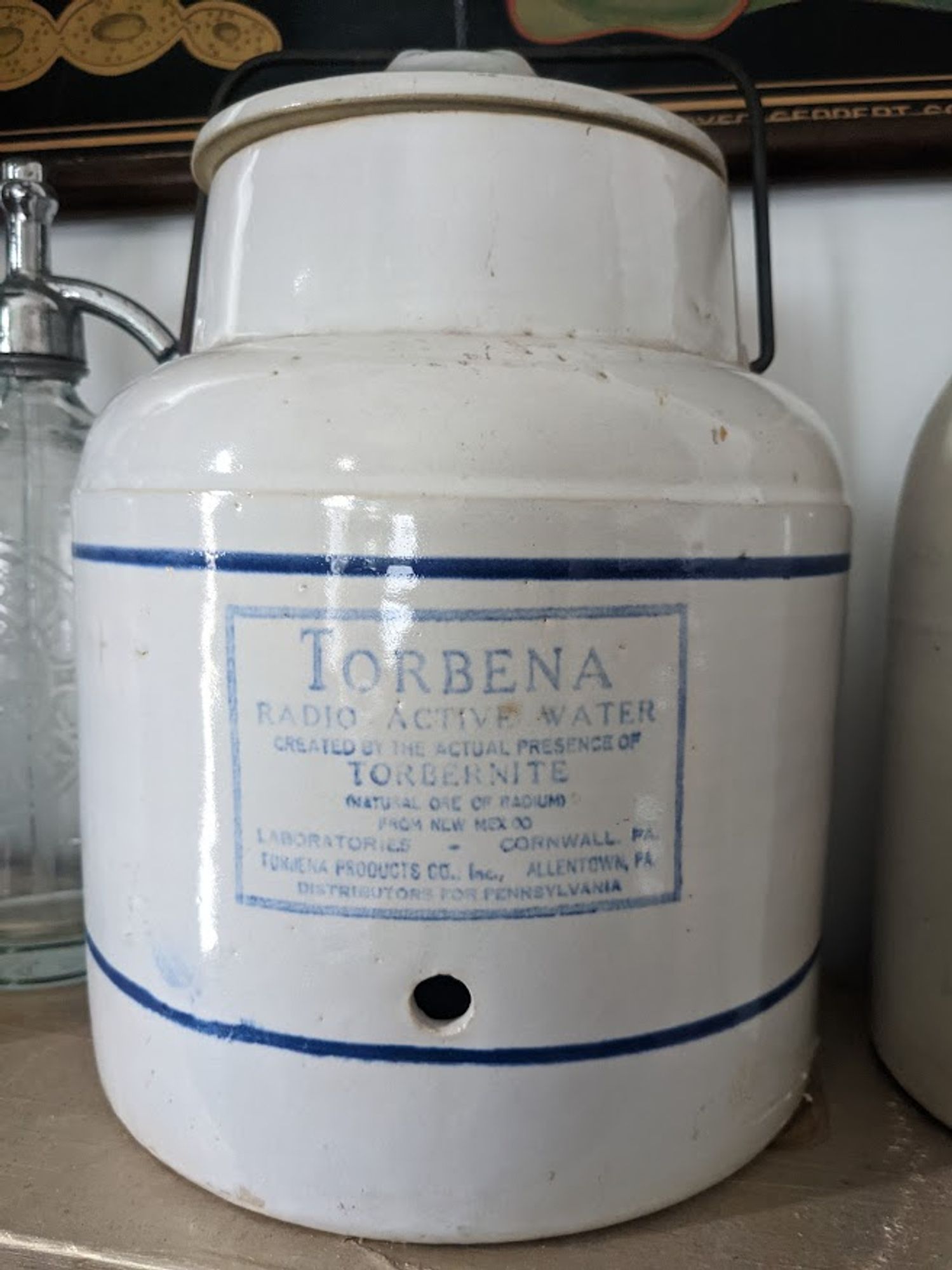 a large white pottery vessel with lid, stamped with an blue label identifying it as once containing Torbena radioactive water "created by the actual presence of Torbenite"