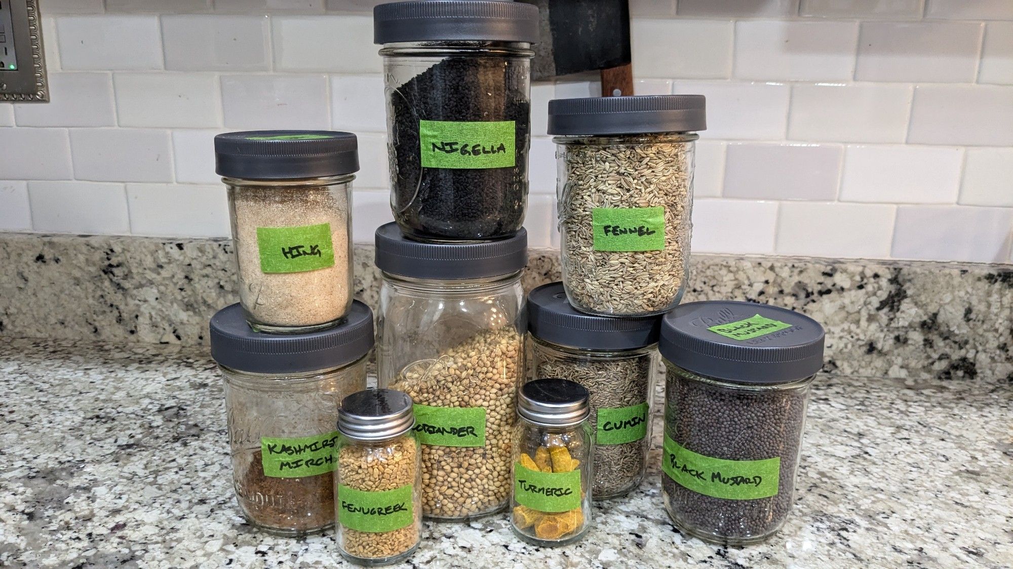 A bunch of mason jars and spice jars, all filled with whole spices, and labeled with sharpie on green tape: hing, nigella, fennel, kashmiri mirch, fenugreek, coriander, turmeric, cumin, black mustard