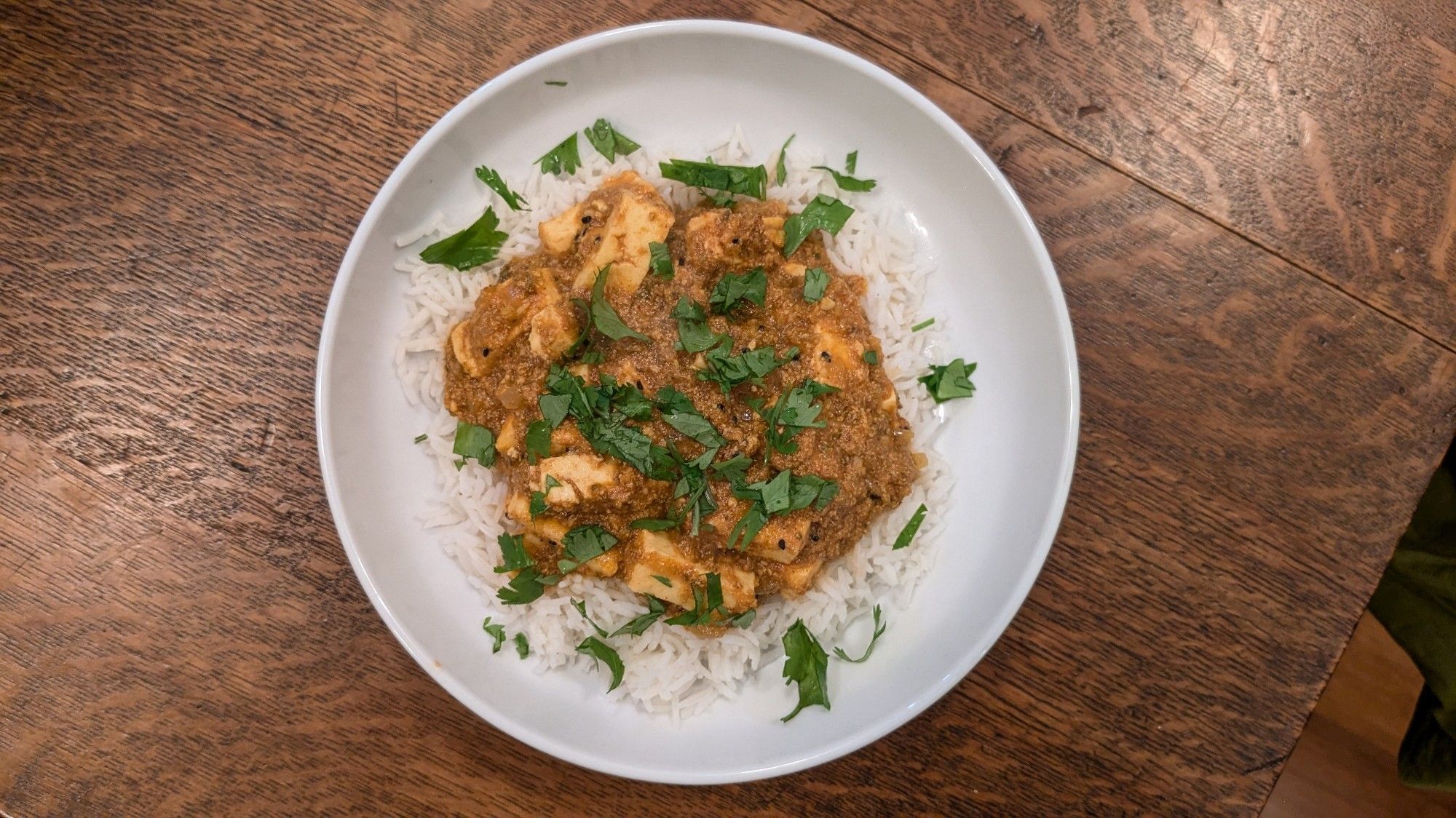 A curry with paneer, on rice, in a white bowl on a wood table, sprinkled with coriander leaves