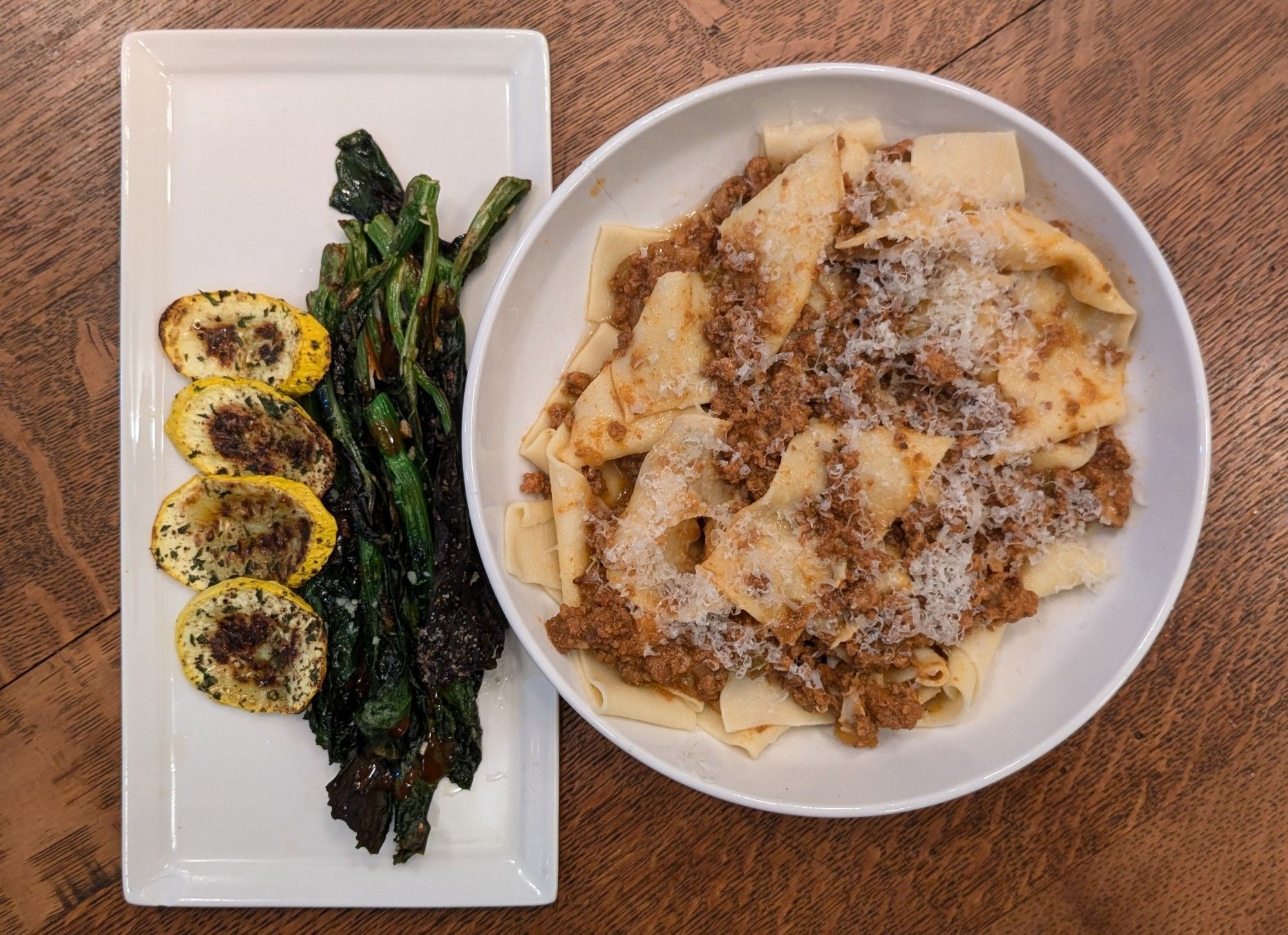 A shallow bowl of wide pasta with ragu and parm, accompanied by a rectangular plate of charred Chinese broccoli tips and yellow summer squash slices.