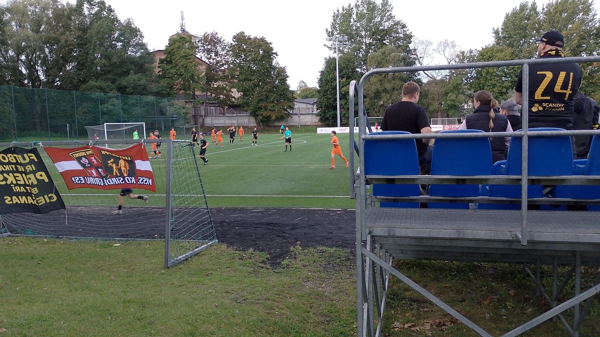 Links ein Tor mit Fahnen, rechts eine Stahlrohrtribüne mit Sitzschalen. Dahinter ein Fußballplatz