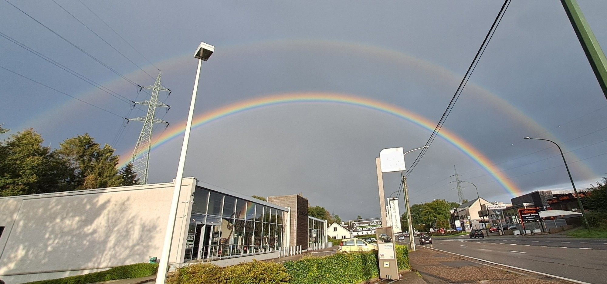 A double rainbow on a grey sky. In the front, a road, buildings, signs and street lamps