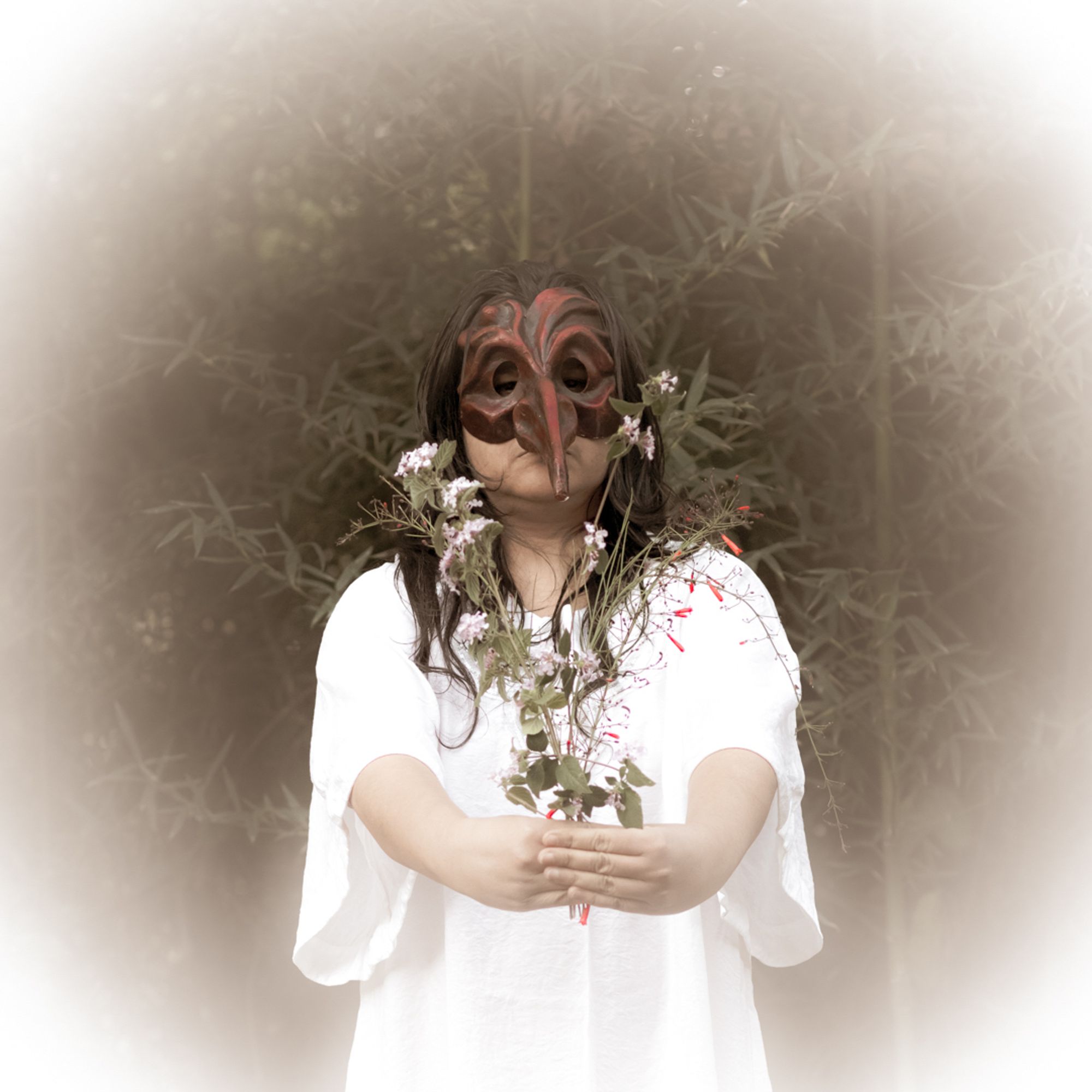 Foto colorida de uma mulher morena com um vestido branco e uma máscara vermelha. Ela segura com os braços esticados alguns galhos com flores. No fundo galhos de bambu verde. Uma vinheta branca contorna a foto.