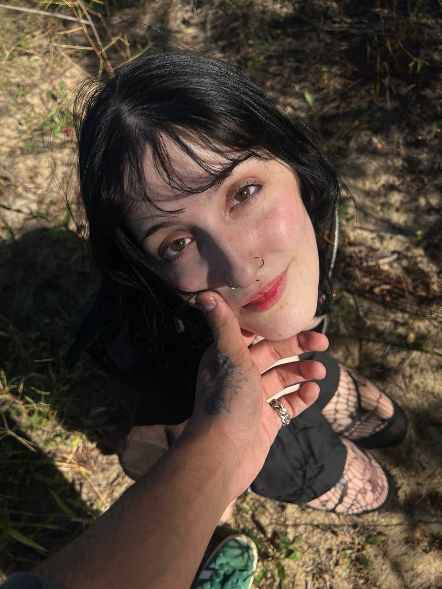 Goth girl looks up at camera. She is wearing a black mini skirt and fishnets with a brown jacket. Her black hair is long with bangs. Her brown eyes are illuminated by the sun, her lips full. A tattooed hand grasps her chin softly , tilting her head upward. 