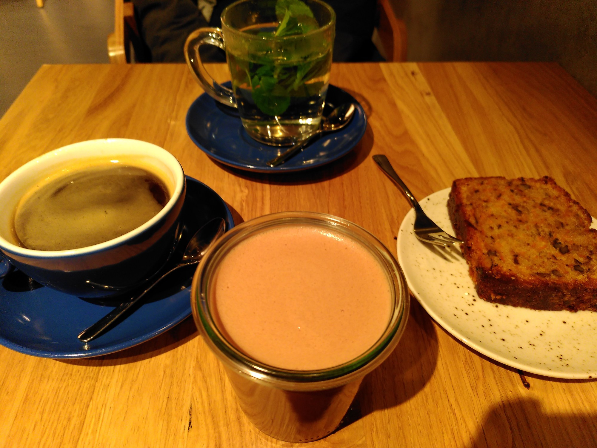 Eine Tasse  Kaffee, ein Pfefferminztee und eine heiße Schokolade mit Hafermilch. Dazu ein Stück Karottenkuchen mit Zimt und Kardamom 