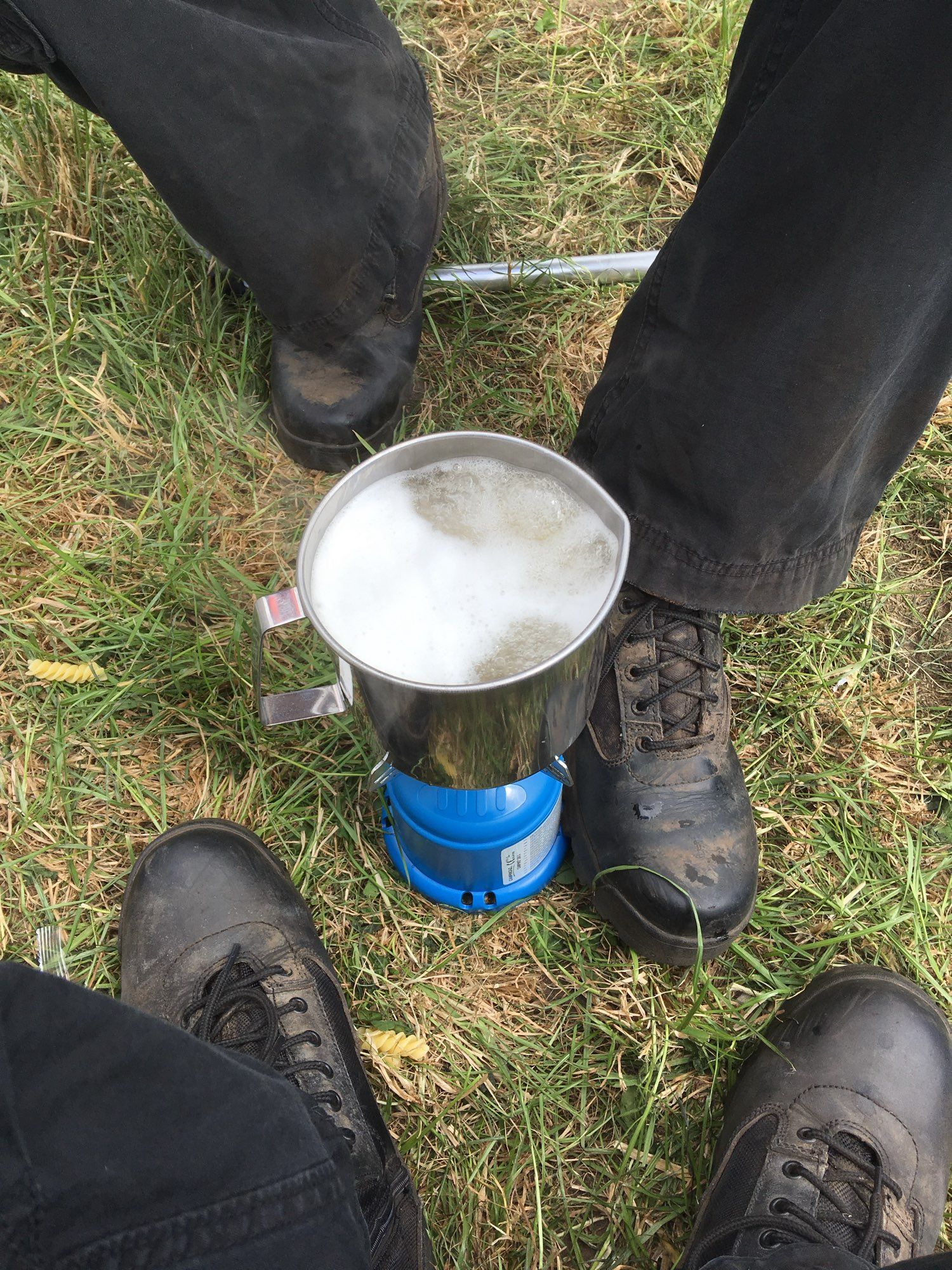 Auf einer Wiese steht ein kleiner Gaskocher mit einem Topf Wasser drauf. Das Wasser kocht. Um den Kocher sieht man schwarze Stiefel von uns, als wir drauf warten, dass die Nudeln fertig werden. Aufgenommen in Belgien auf dem Graspop Metal Meeting