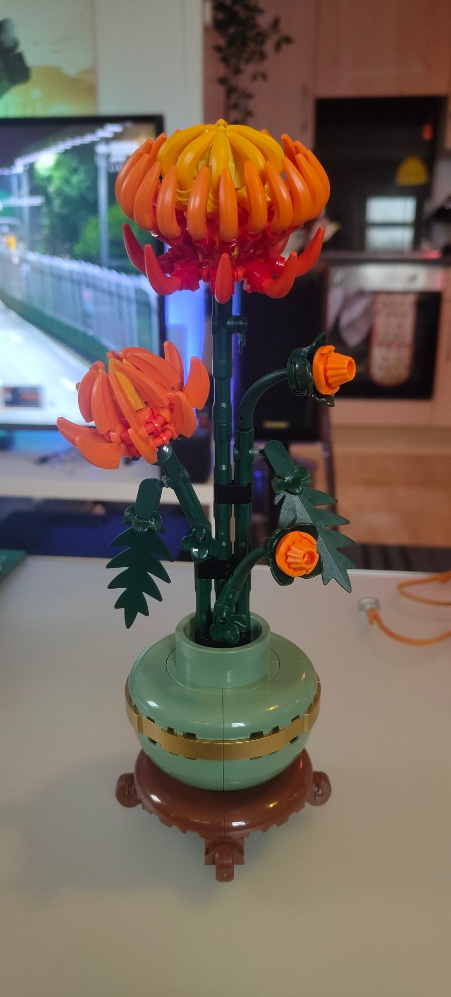 A chrysanthemum made out of Lego. It stands in a jade green and brown vase with a gold band, also Lego. Three green stalks are topped by yellow-orange blossoms, made mostly out of Lego banana pieces.